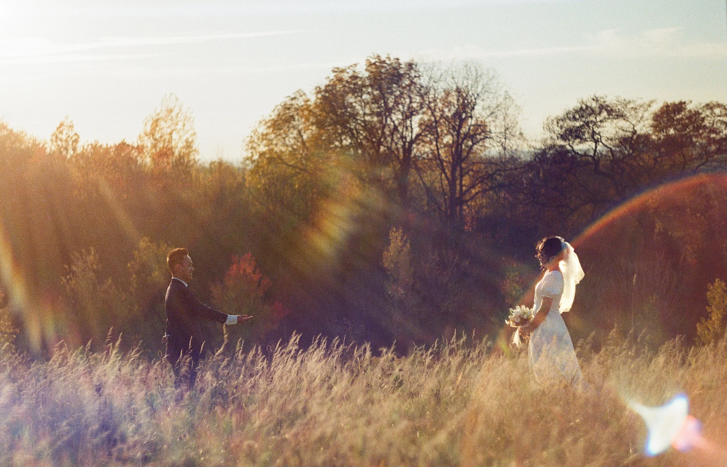 A bride and groom in a field during sunset, with the groom reaching out to the bride who holds a bouquet, surrounded by tall grass and autumn-colored trees.