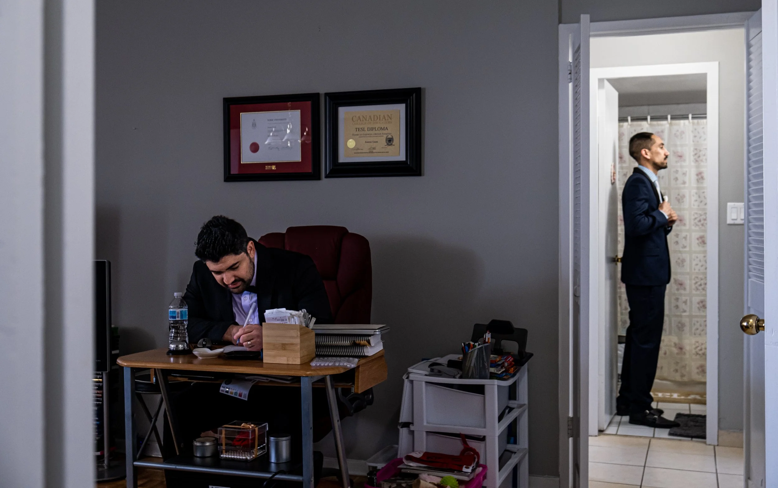 An office with a man sitting at a desk and another man standing in a bathroom doorway. The man at the desk is looking down, wearing a black blazer, with documents, a water bottle, and office supplies on, and framed diplomas on the wall behind him. Th
