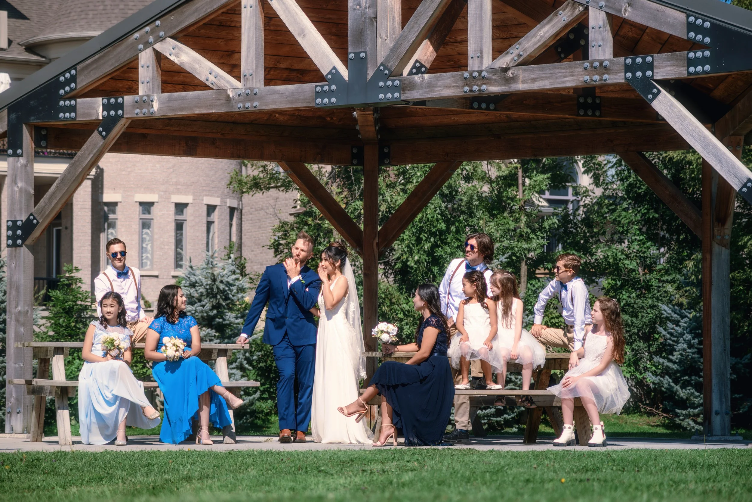 A wedding ceremony taking place outdoors under a wooden pavilion, with a bride in a white dress and a groom in a blue suit standing in front of guests, including flower girls and page boys.