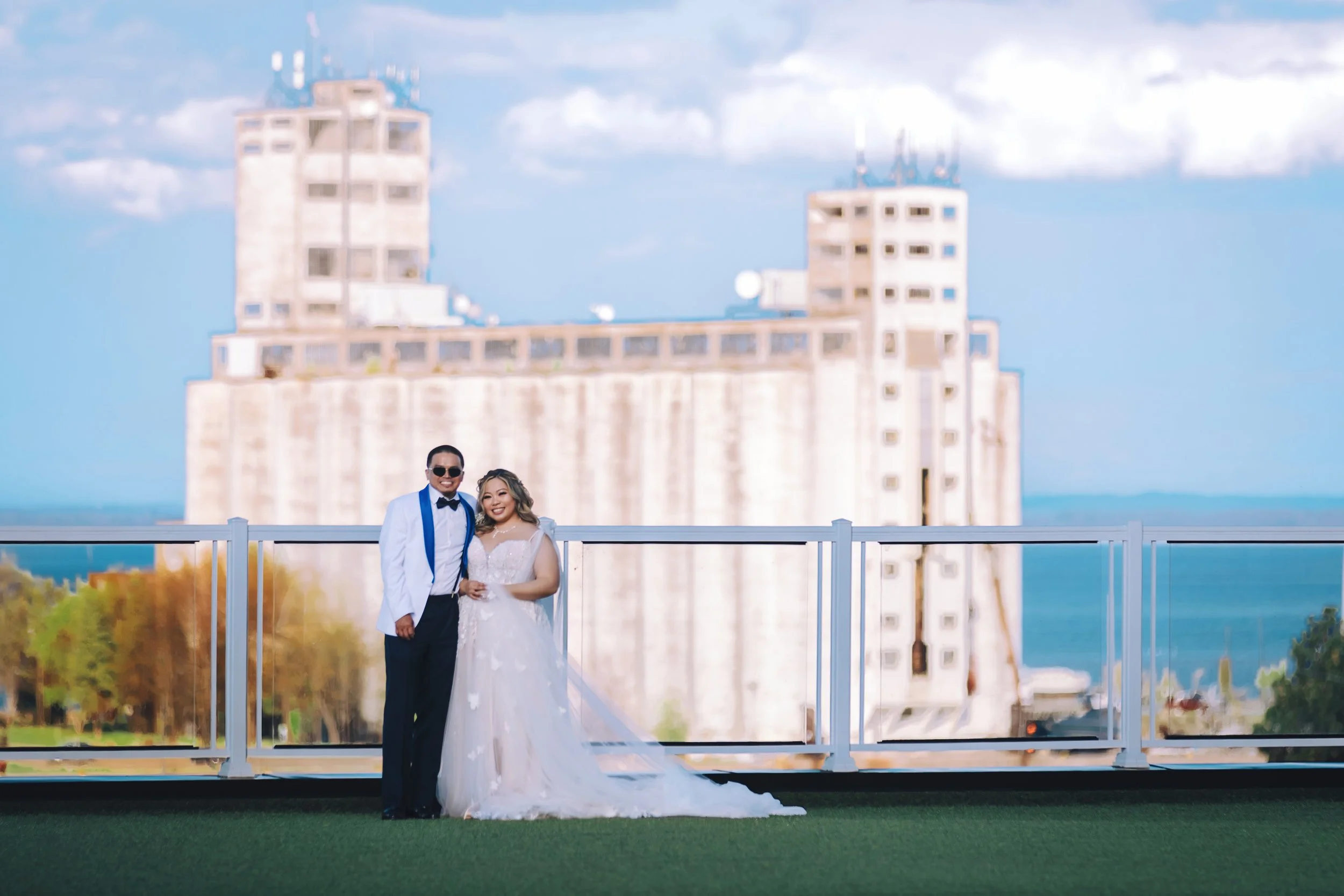 A newlywed couple dressed in wedding attire, standing outdoors on a rooftop with a city skyline and water in the background.