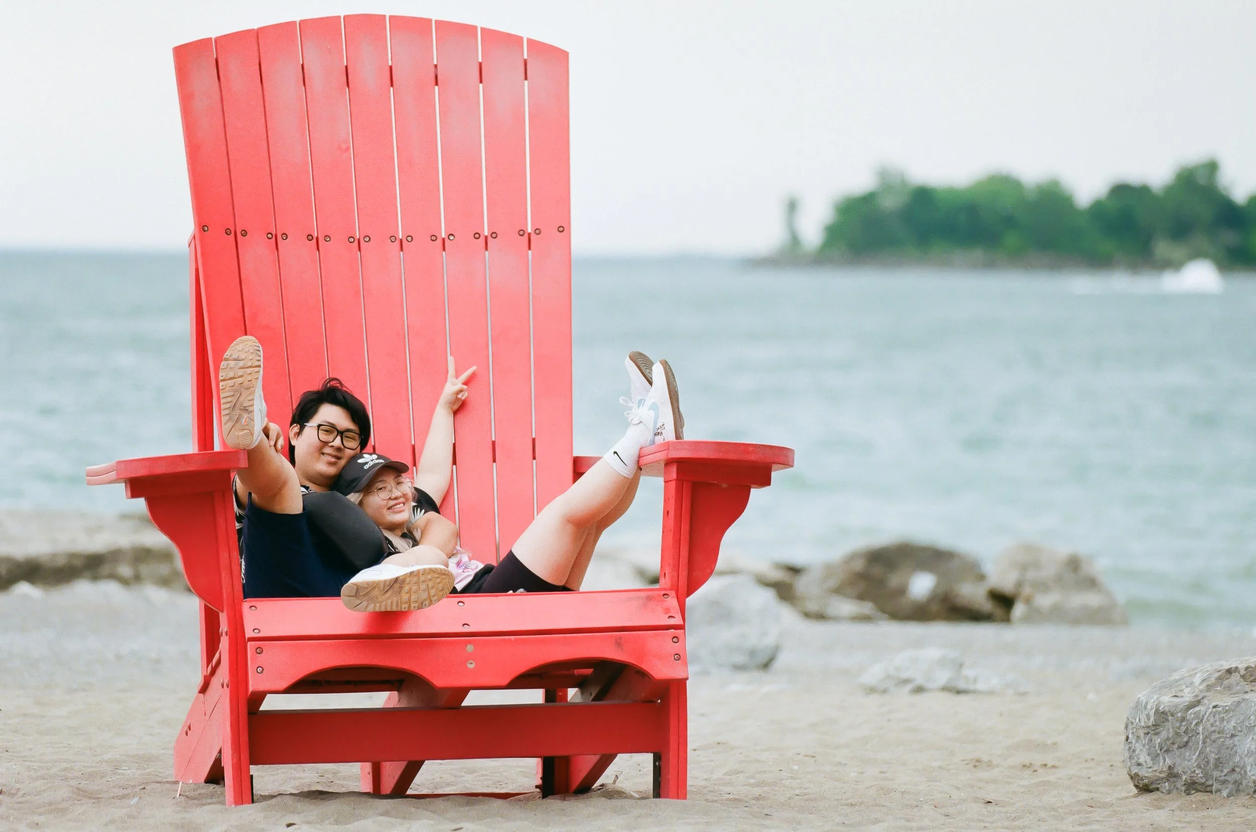 Two people sitting on a large red Adirondack chair on a beach, smiling and relaxing by the water, with rocks and a distant tree-lined shoreline in the background.