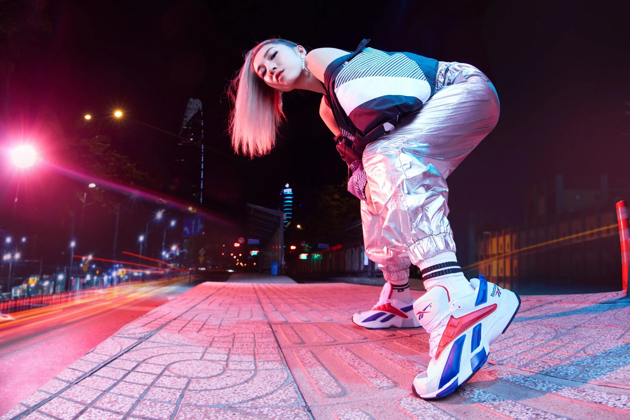 A young woman in reflective silver pants, a striped jacket, and Reebok sneakers is crouching on an urban sidewalk at night with city lights and blurred car streaks in the background.