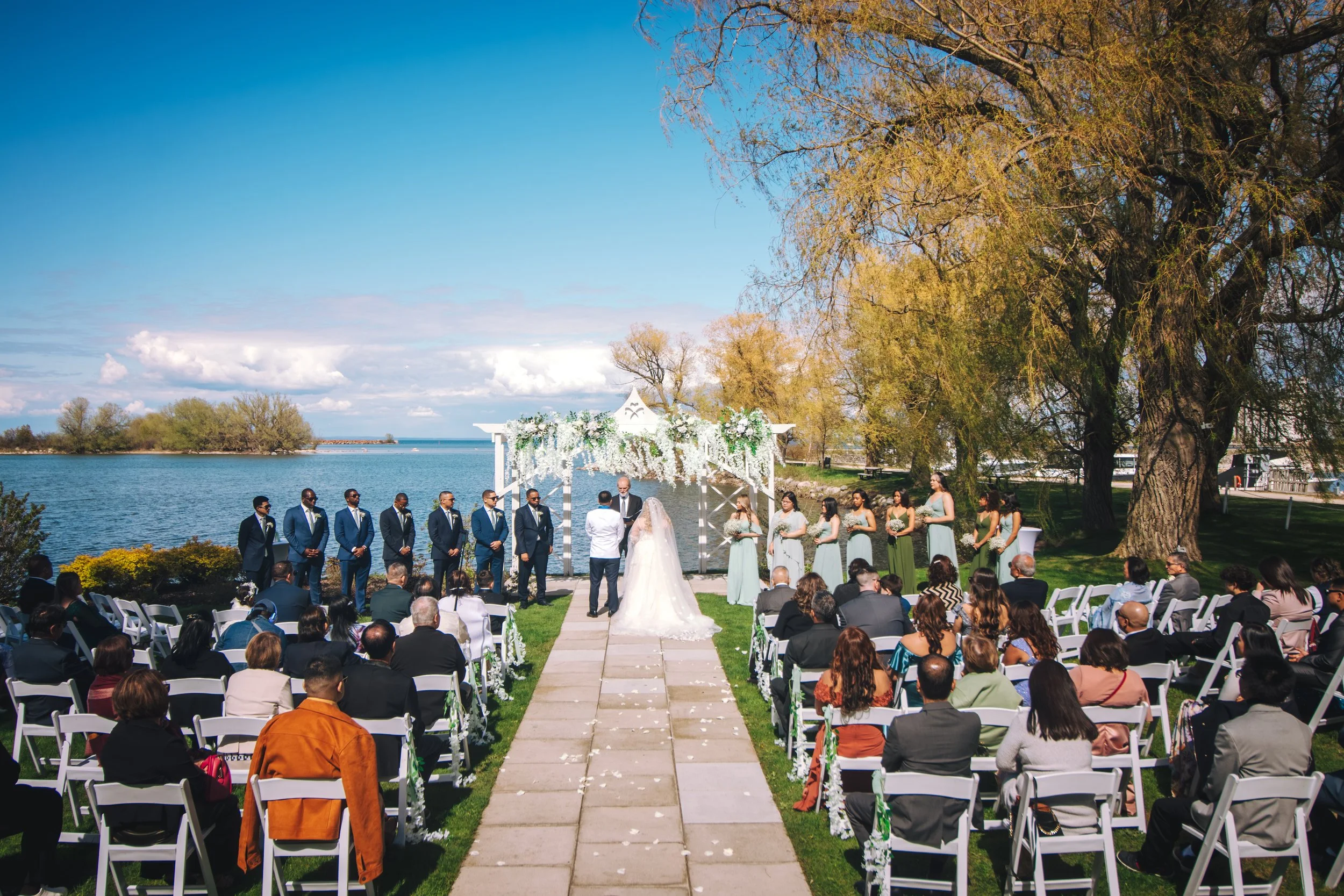 Outdoor wedding ceremony by a lake with guests seated on white chairs, a white wedding arch decorated with white flowers, a bride and groom exchanging vows, surrounded by bridesmaids and groomsmen, large trees and a blue sky in the background.