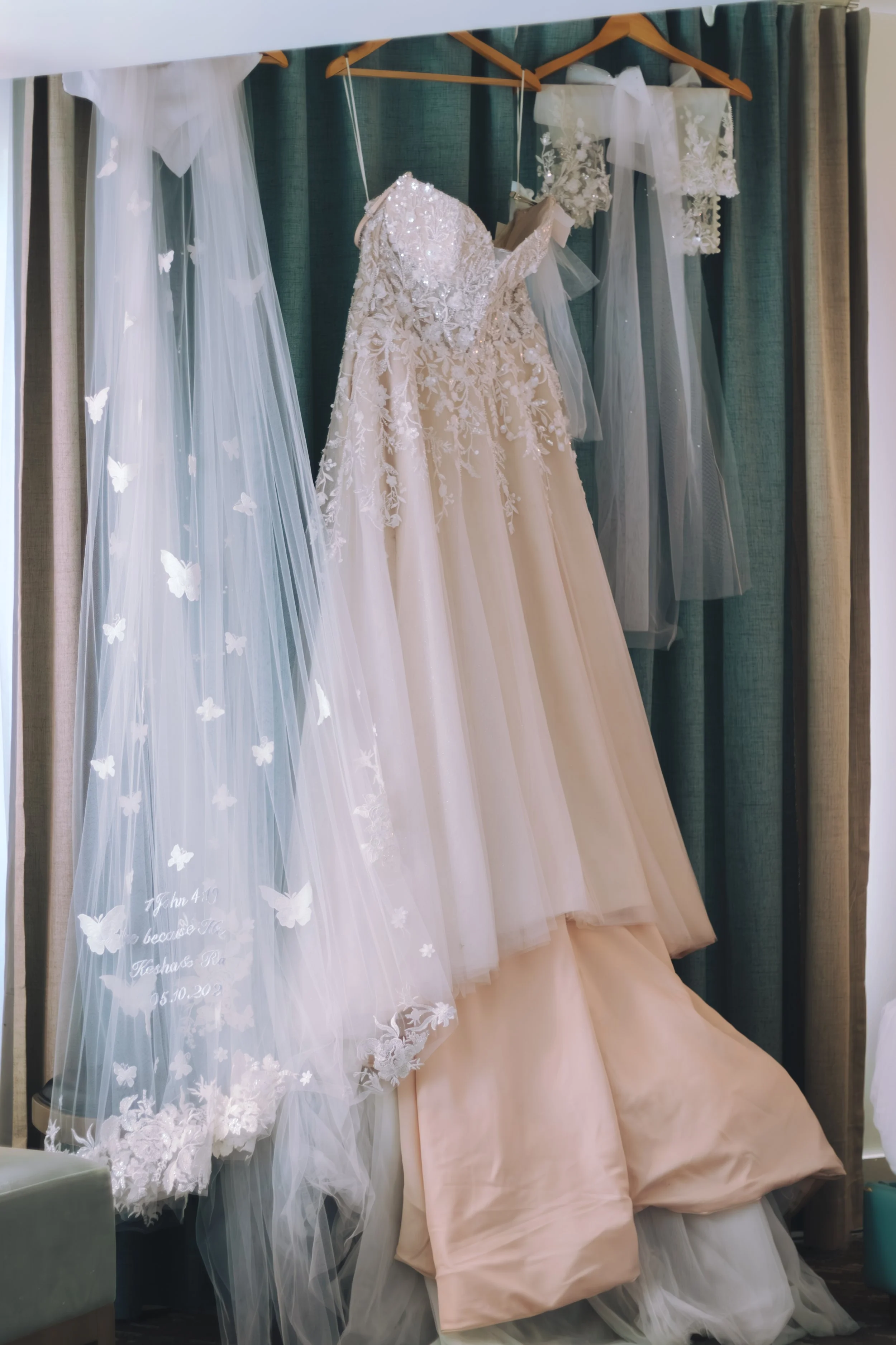 A wedding dress with lace and beaded details hanging on a wooden hanger, accompanied by a sheer veil with lace appliqué and butterfly embroidery, also hanging on a hanger, with curtains in the background.