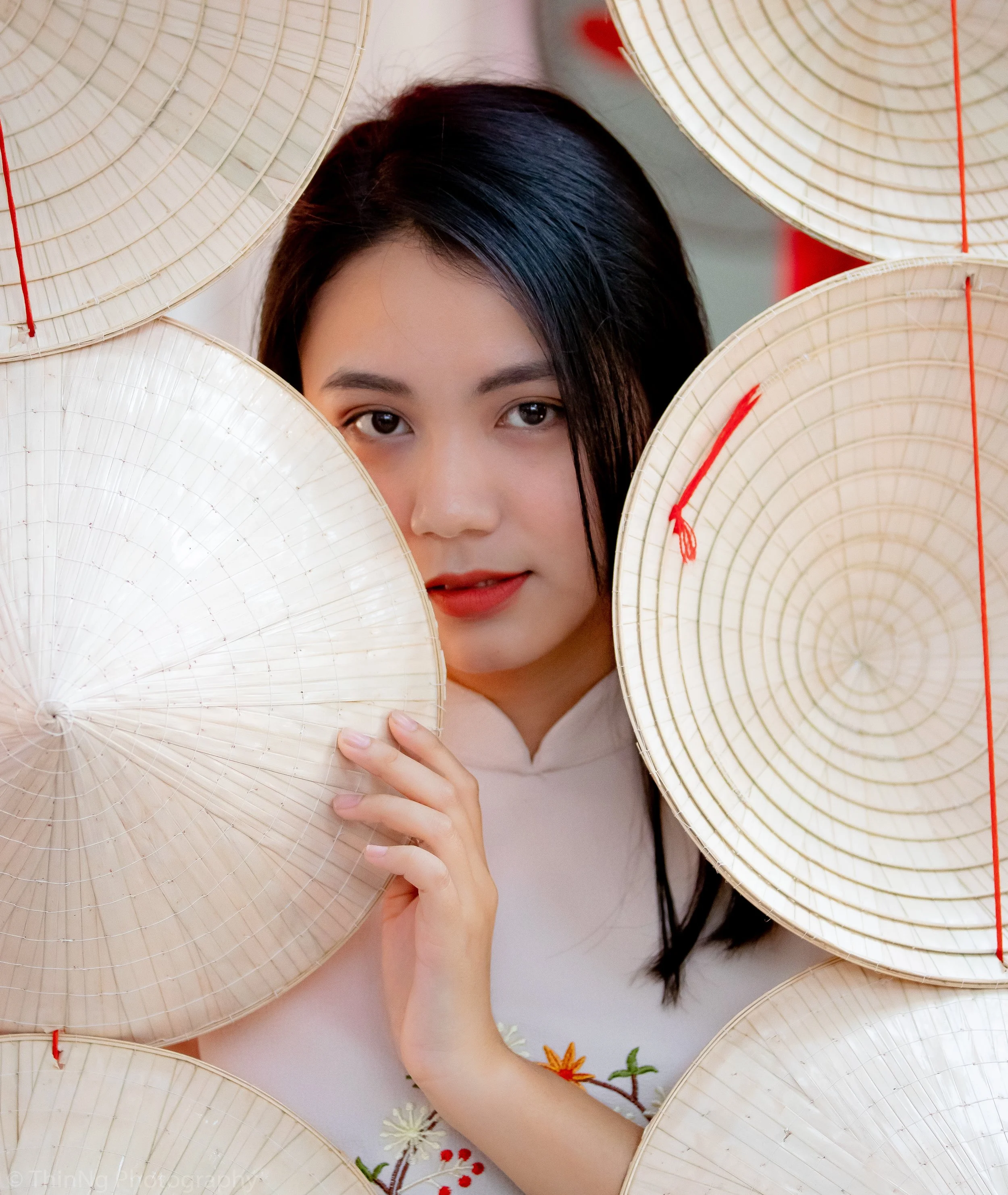 A woman with dark hair and makeup peeking through a group of white Asian conical hats with red strings.
