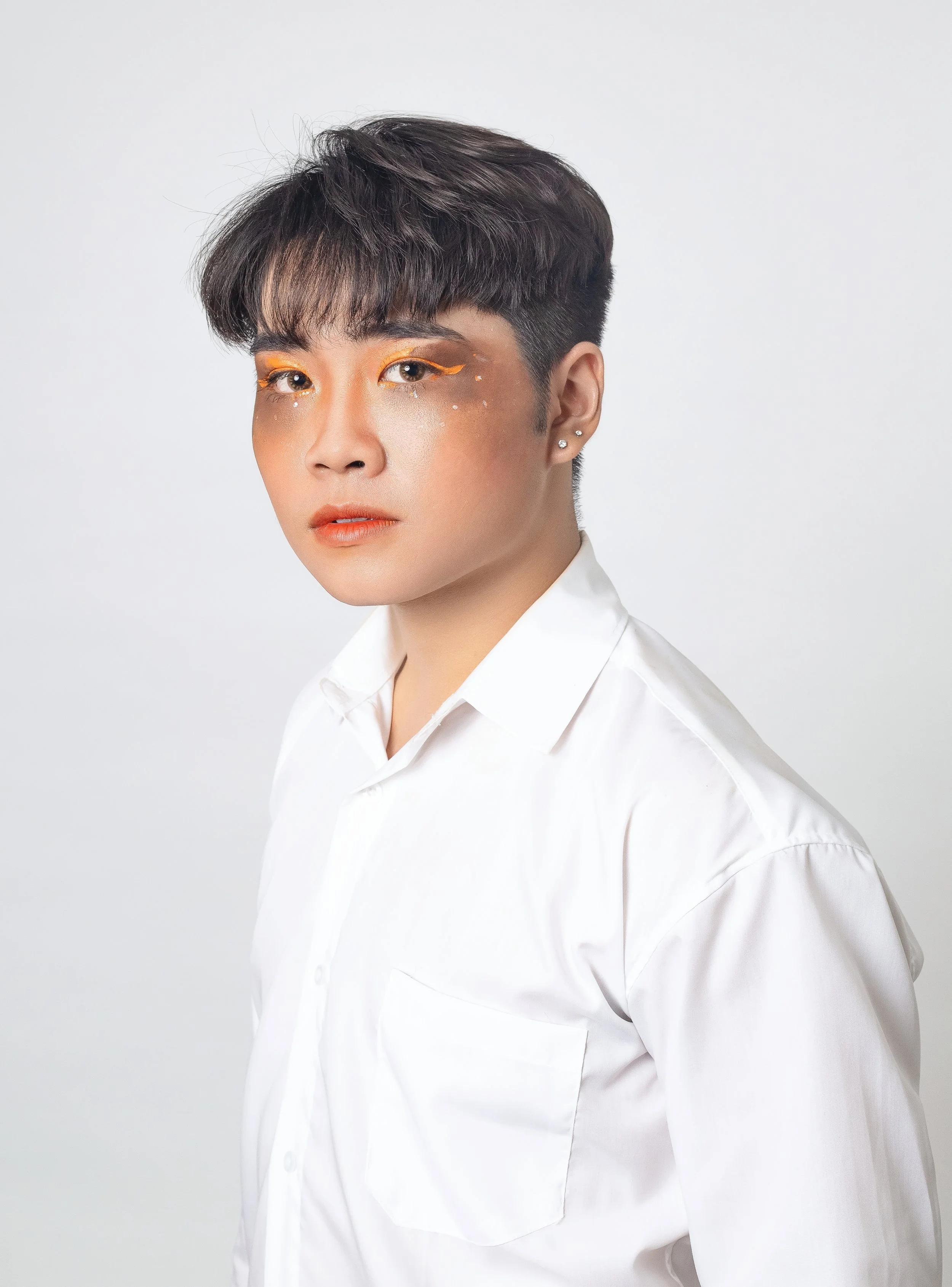 A person with short dark hair wearing a white shirt with makeup featuring yellow eyeliner and decorative rhinestones around the eyes, posed against a plain light gray background.
