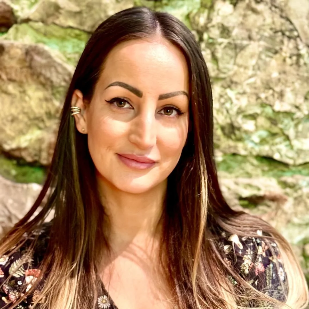 A woman with long brown hair and light skin, smiling outdoors in front of a rocky background with greenery.