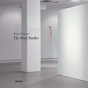 An art gallery with white walls; a floating mannequin in beige clothing near the corner. Text on the wall reads 'Werner Reiter The Mind Hacker'.