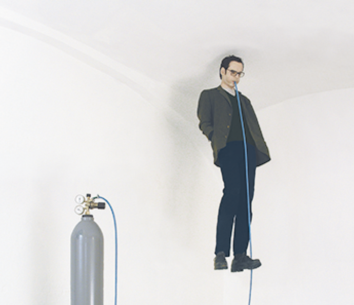A man wearing glasses and a jacket standing on a platform connected to an oxygen tank with a blue tube.