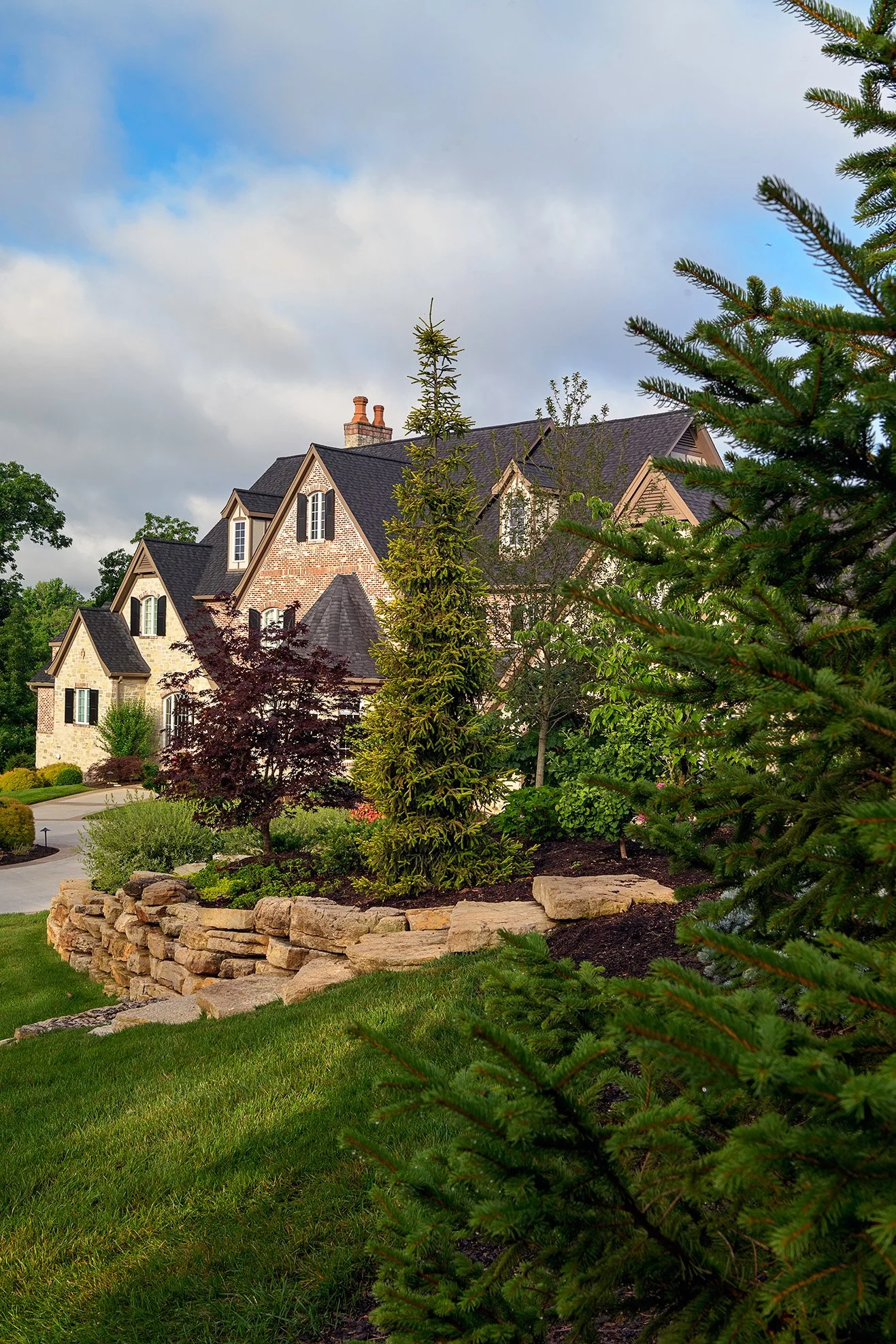 Garden photography of a large house with brick and stone exterior, multiple gabled roofs, and tall chimneys, surrounded by landscaped garden with various trees and shrubs, including conifers, under a partly cloudy sky.