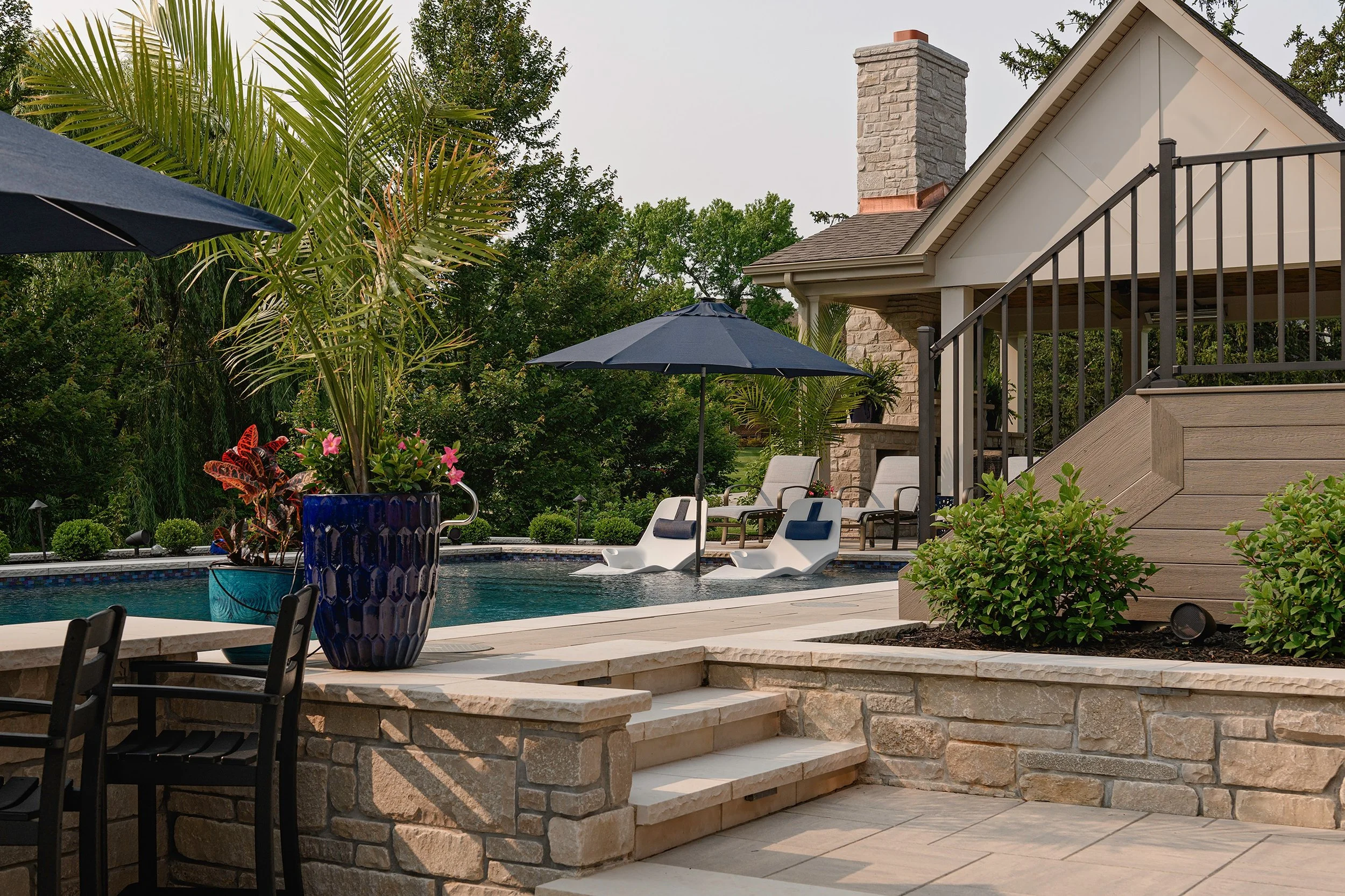 Backyard with swimming pool, blue umbrella, lounge chairs, potted plants, trees, and a house with stone chimney.