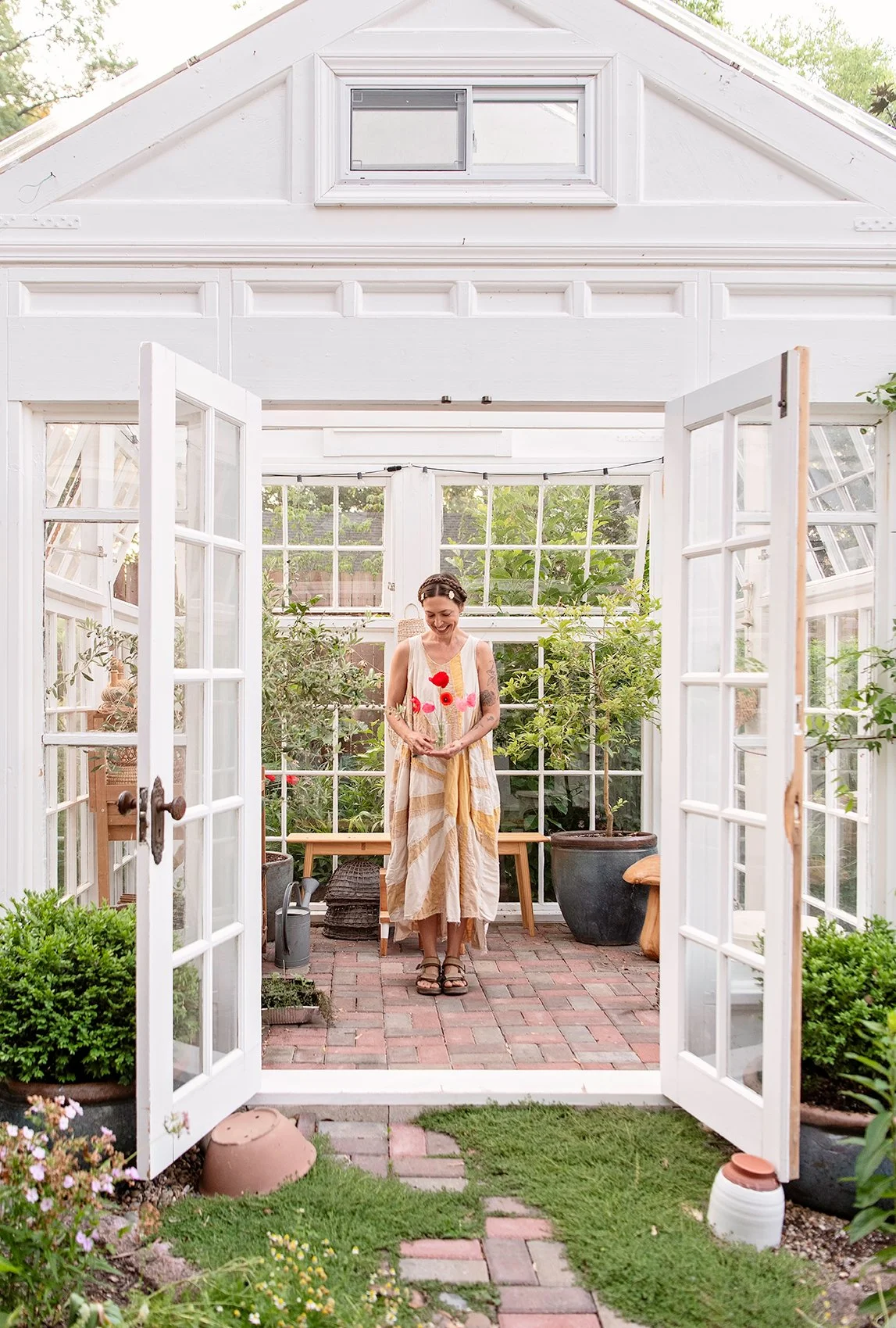 A woman standing in a bright greenhouse with open French doors, surrounded by lush greenery and potted plants.