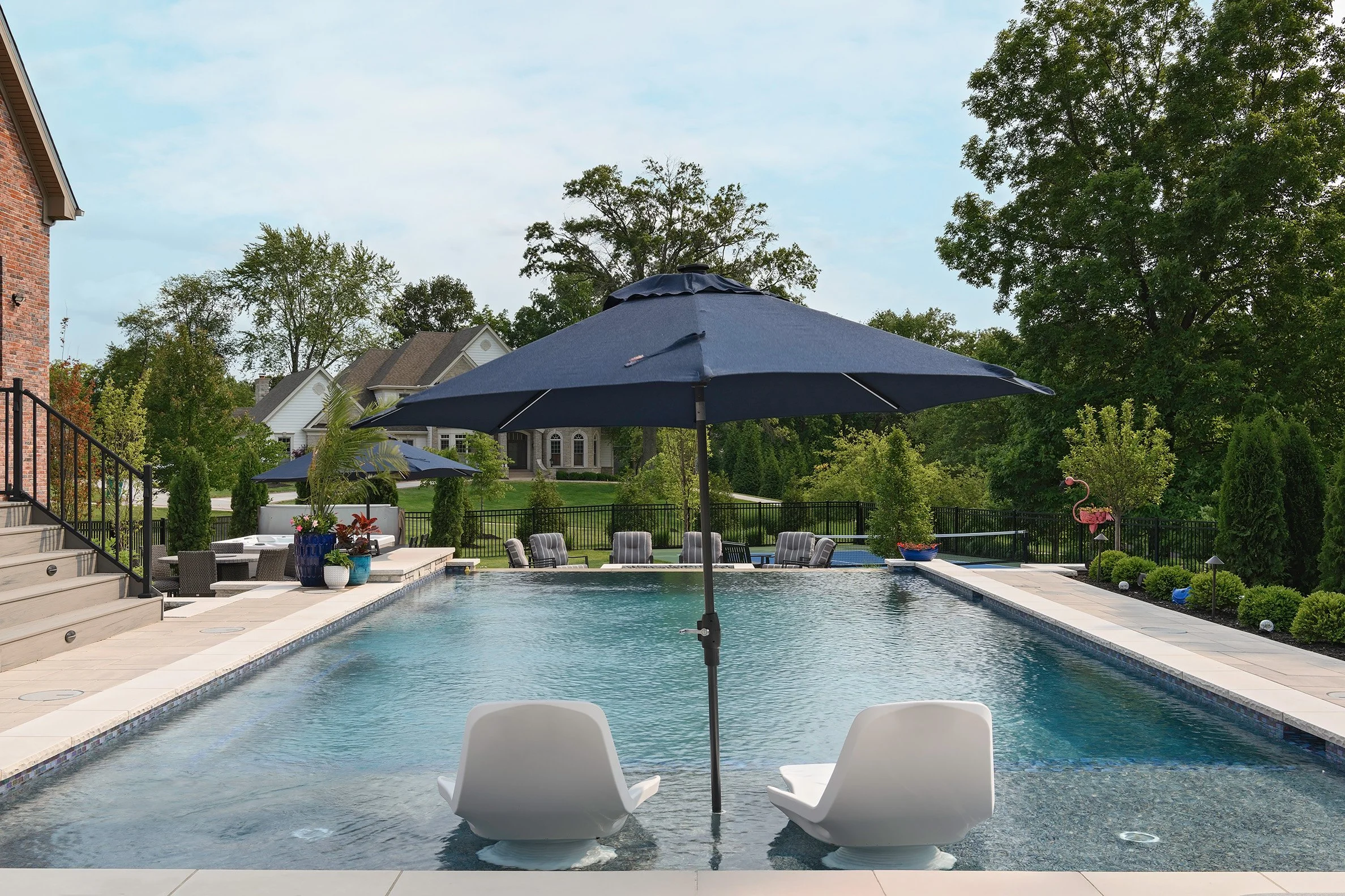 Swimming pool with two white chairs, a blue umbrella, surrounded by outdoor furniture, trees, and houses in the background.