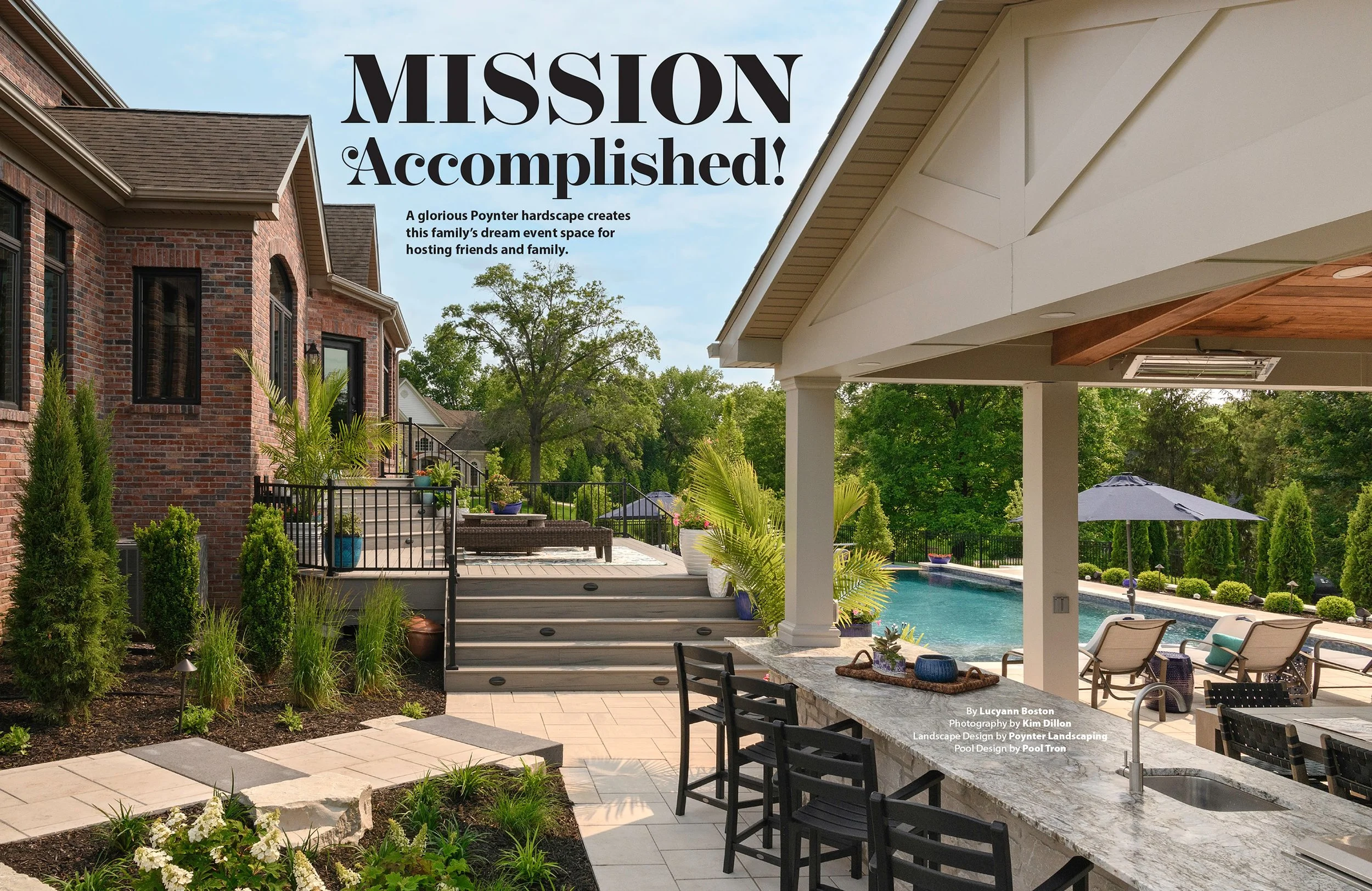 A backyard patio area with a swimming pool, lounge chairs, a shaded bar with barstools, potted plants, lush green trees, and a brick house in the background. The sky is partly cloudy.