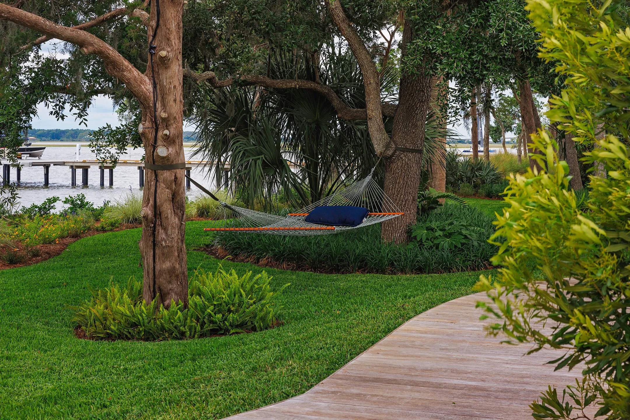 Hammock with a pillow hanging between trees near water, with a wooden walkway and lush greenery in the background.