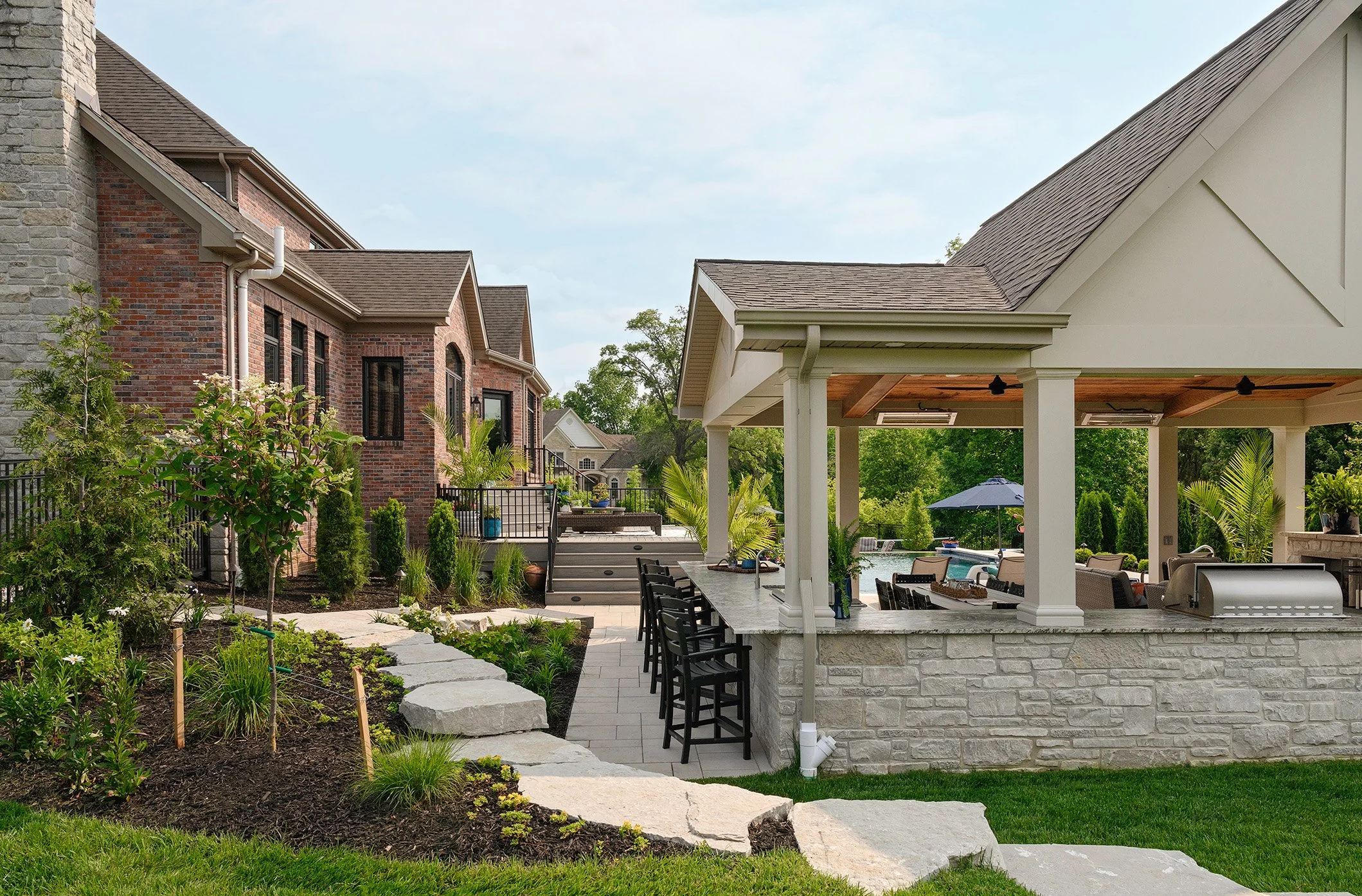 A backyard with a stone patio, bar area with chairs, outdoor grill, garden, and a swimming pool with umbrellas, surrounded by greenery and a two-story brick house. 