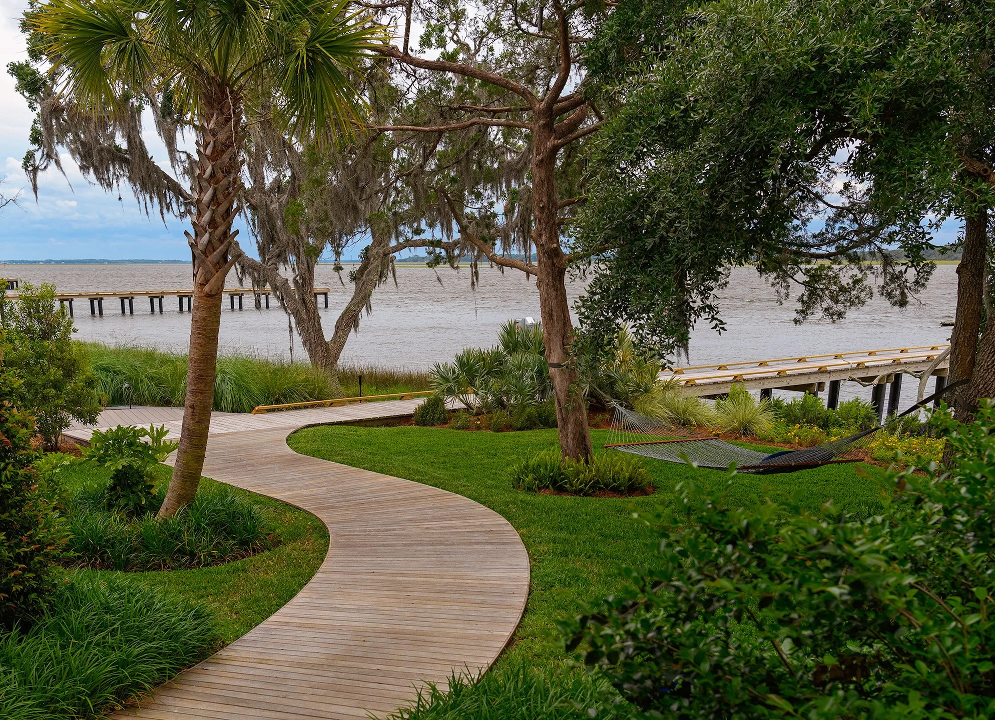 A winding wooden pathway through lush green grass and trees by the water, with hammocks hanging between trees and a pier extending over the water in the background.