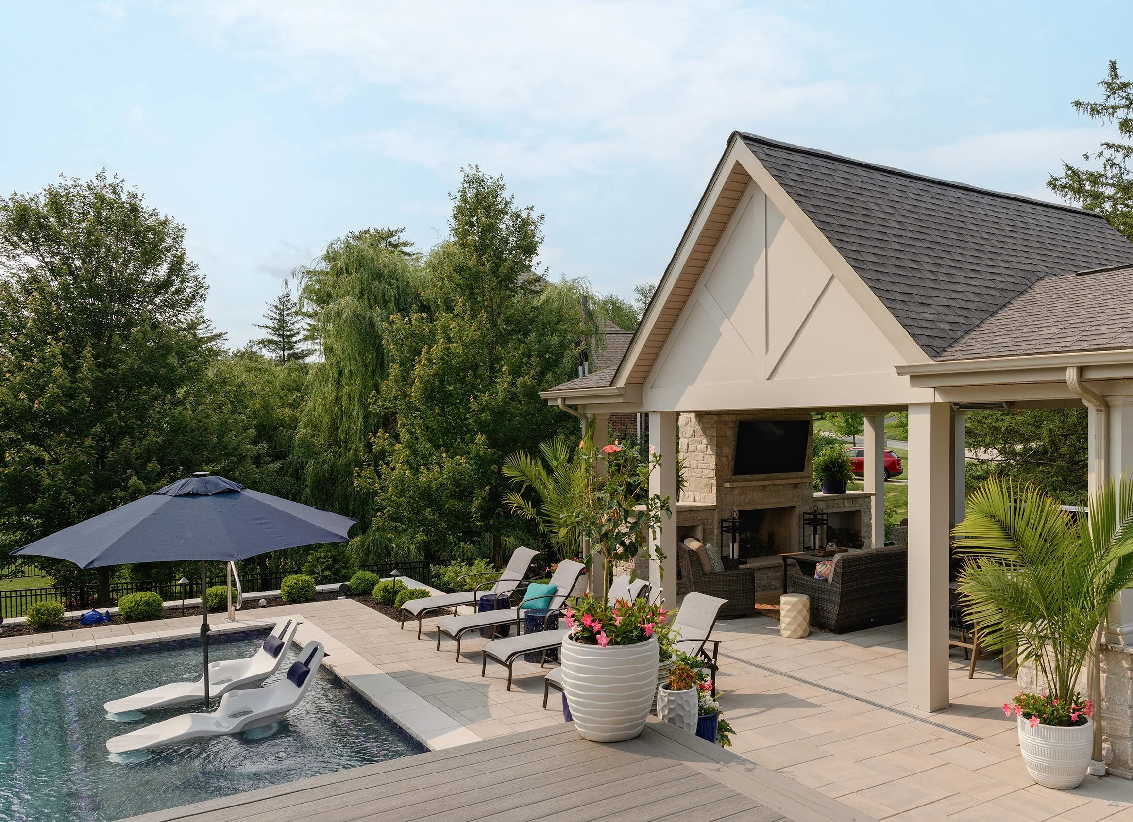 Backyard patio scene with a swimming pool, lounge chairs, umbrellas, potted plants, and an outdoor sitting area under a covered gazebo.