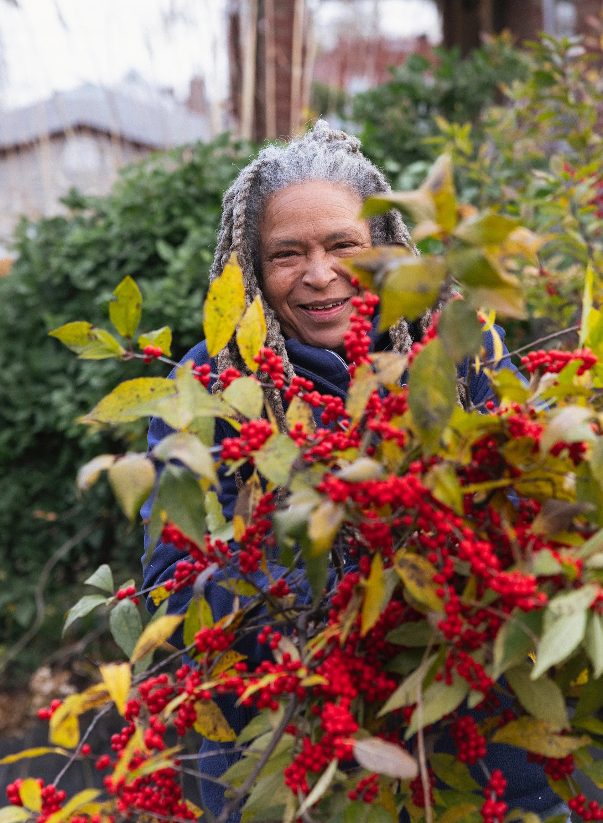 A flower farmer with gray dreadlocks smiling behind a fresh harvest of a red berry bush with green leaves.