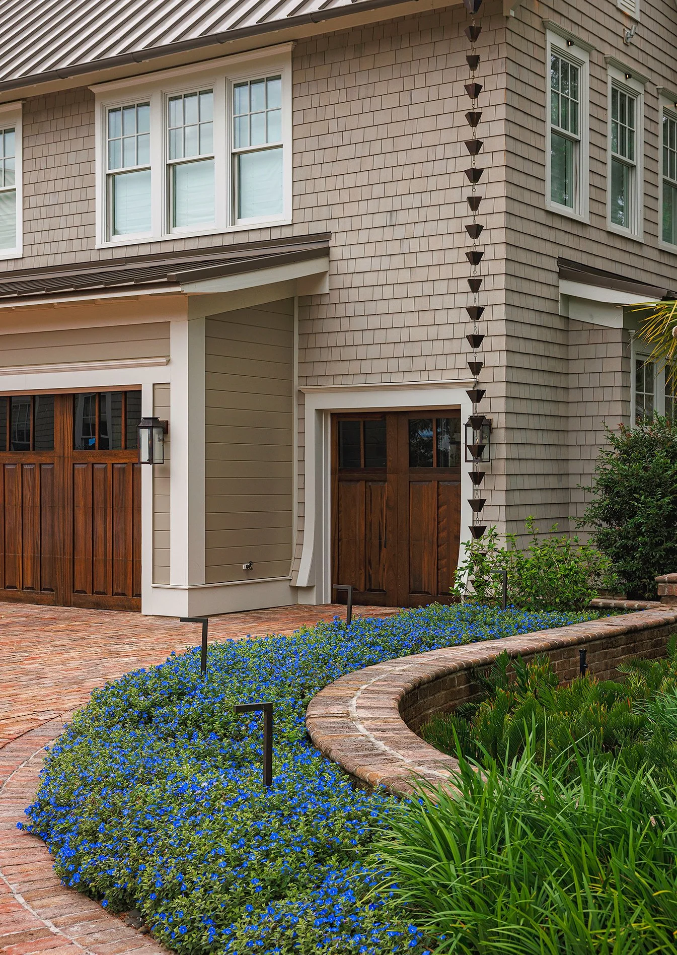 Front view of a house with brick and shingle exterior, wooden garage door, and a small entry door, surrounded by landscaped garden with blue flowers and green plants.