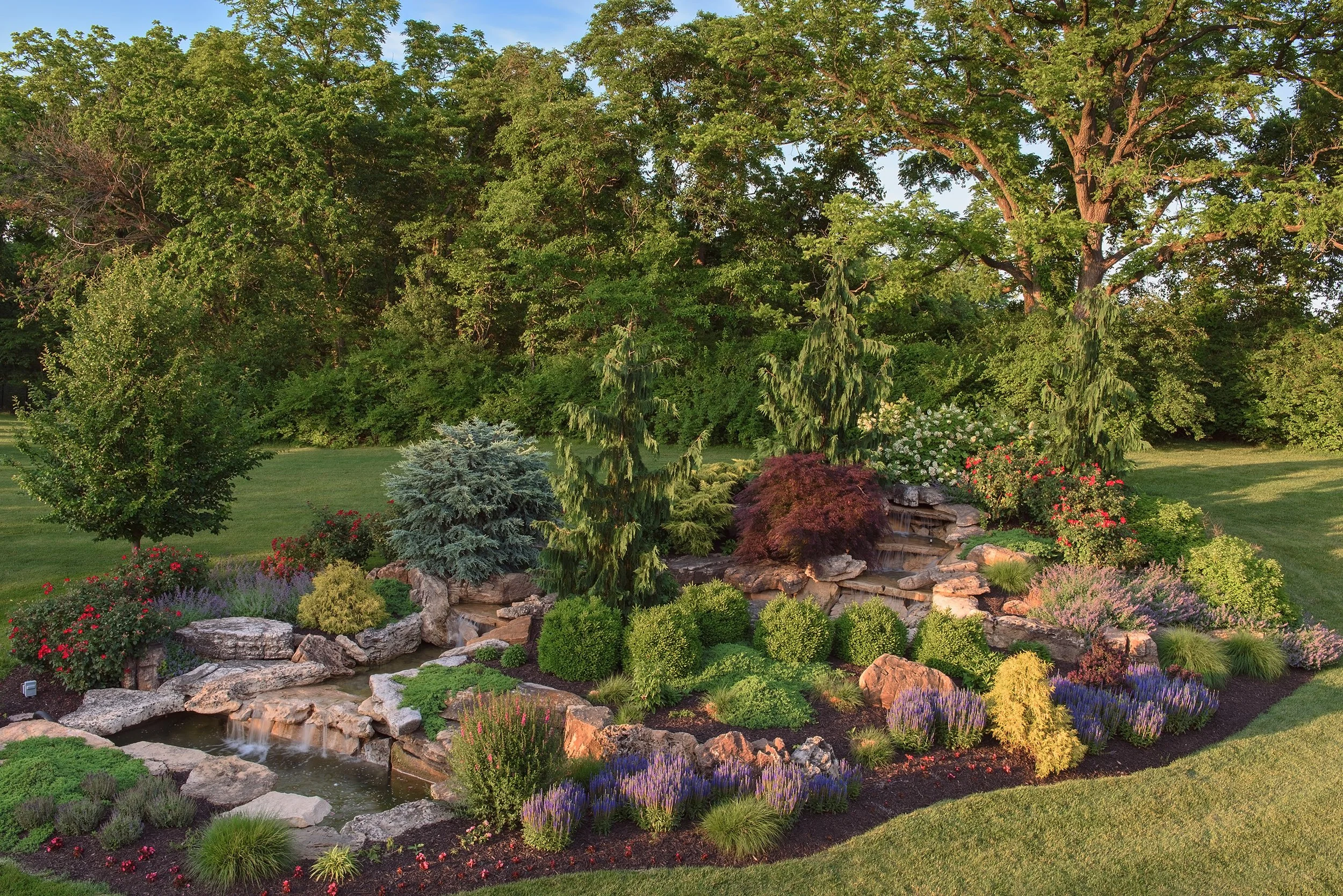 A landscaped garden with a small cascading waterfall surrounded by various green shrubs, flowering plants, and tall trees, set on a well-maintained grassy lawn.