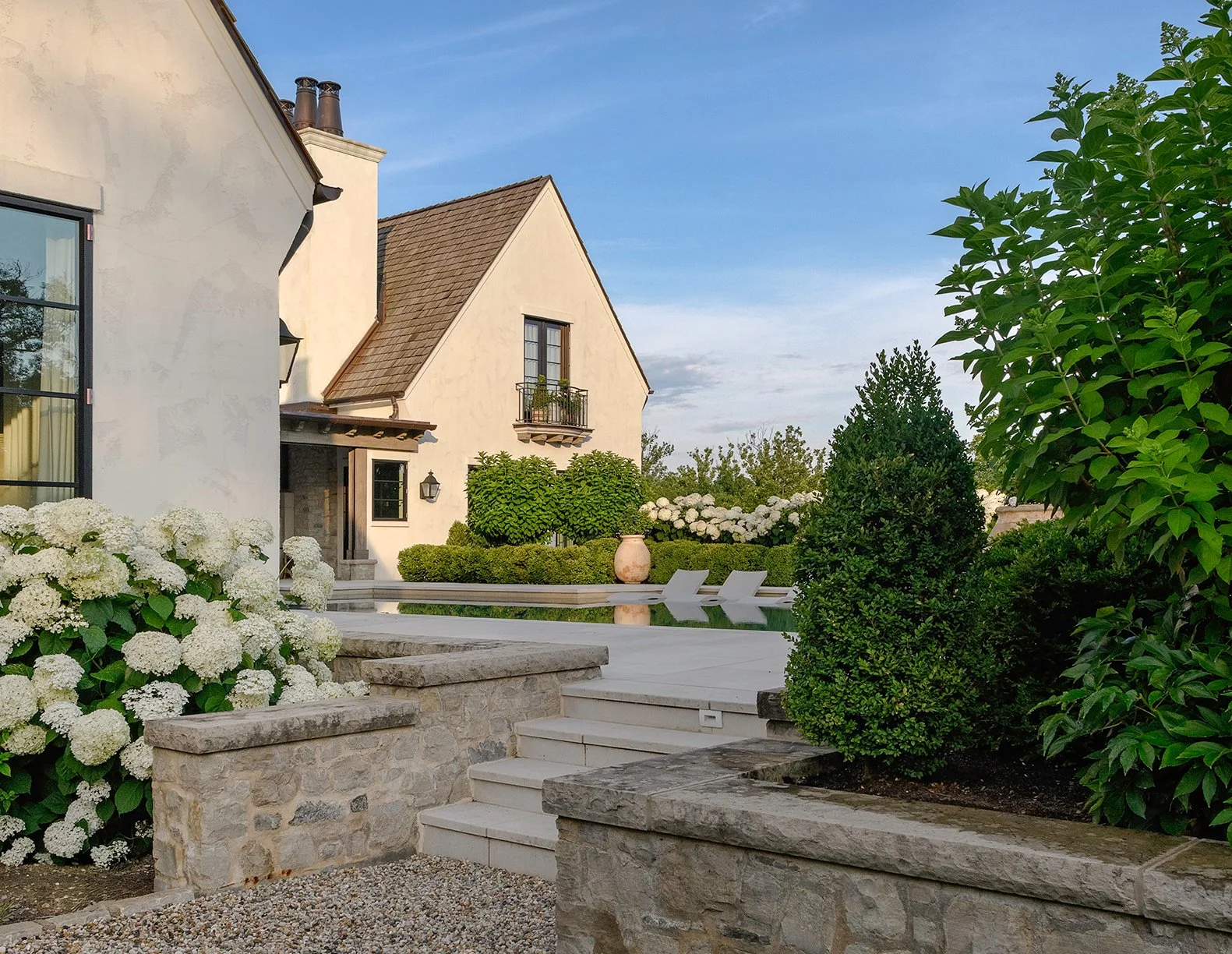 A backyard with a stone patio, white chairs, and lush green bushes and trees, adjacent to a beige house with a gabled roof and balconies, under a partly cloudy sky.