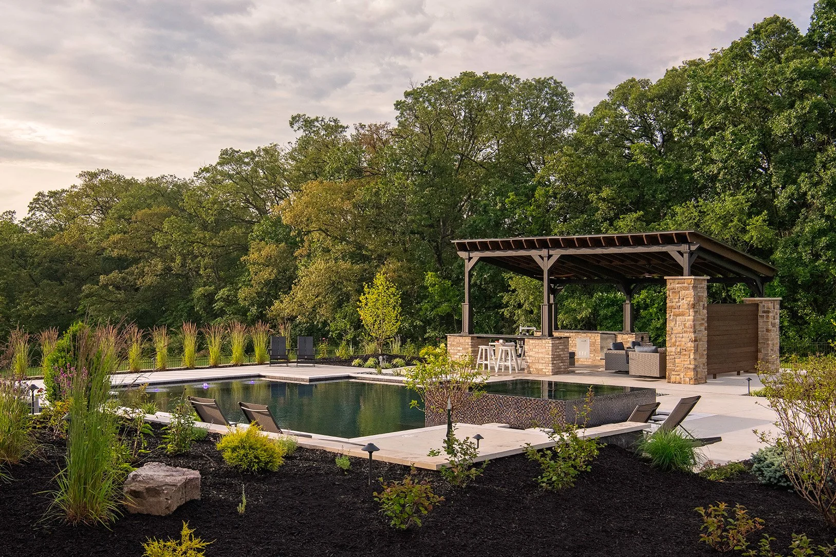A backyard with a swimming pool, a poolside lounging area with chairs and plants, and a covered outdoor kitchen with stone and wood accents, surrounded by lush green trees and a cloudy sky.