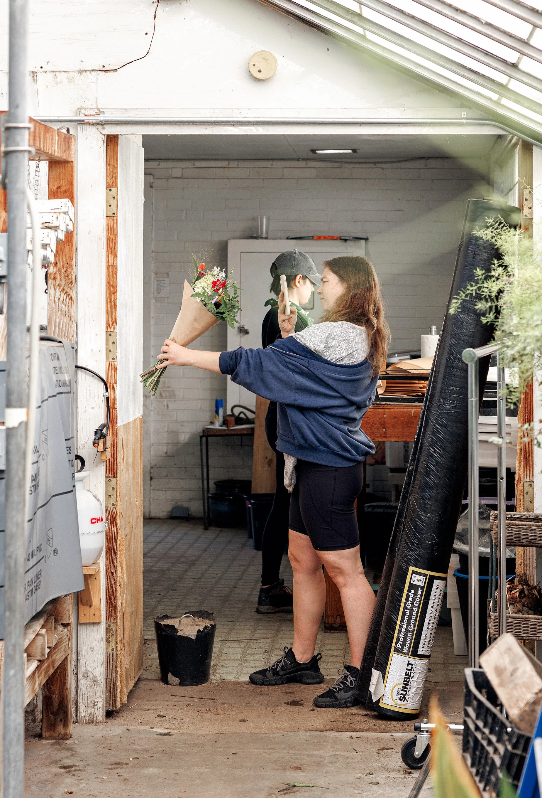 A woman is holding a bouquet of flowers and taking a photo of a young person in a greenhouse or garden workspace with tools and gardening supplies around.