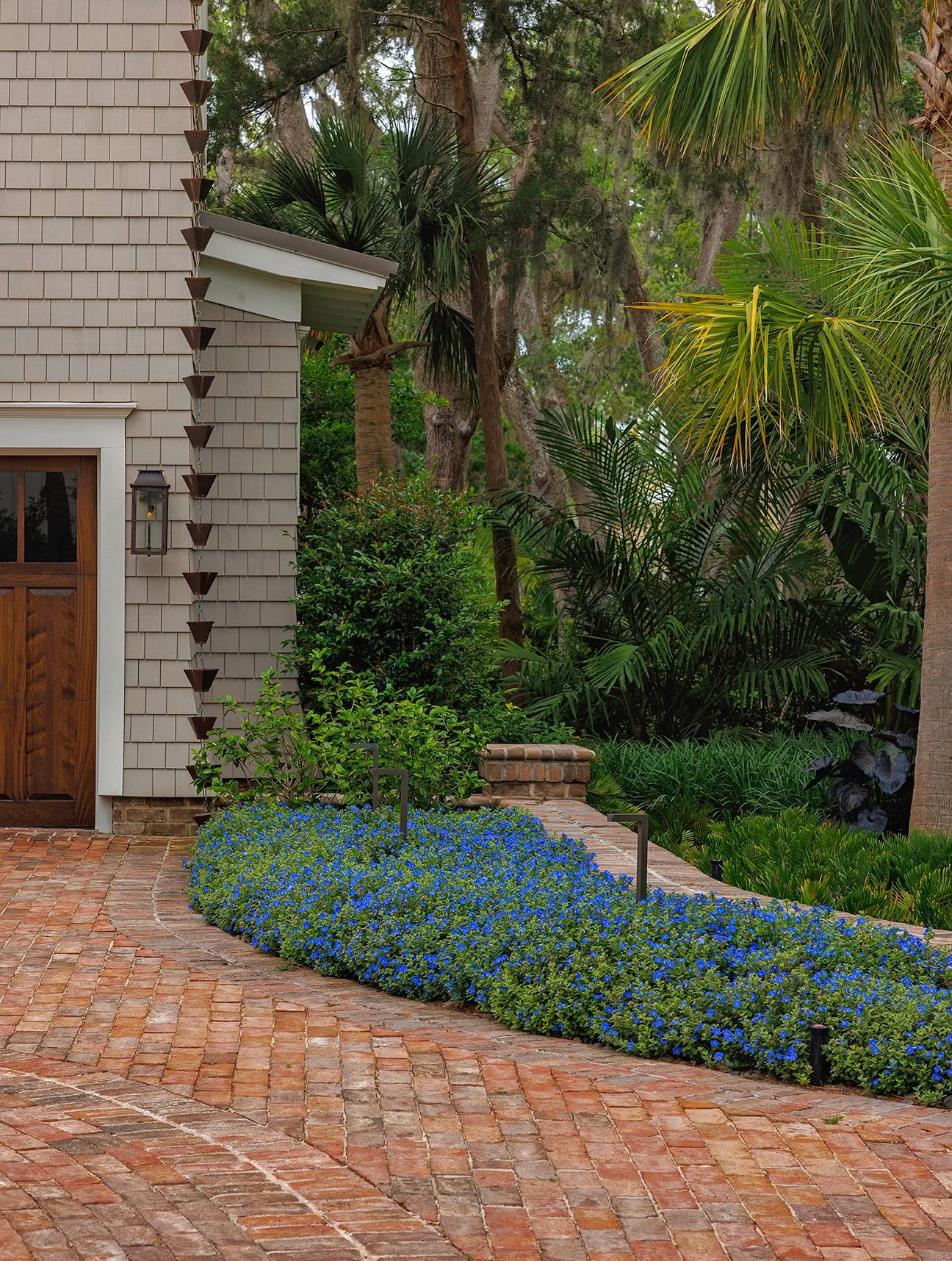 A brick pathway next to a house with beige shingles, green bushes, and a garden with tropical plants and trees.