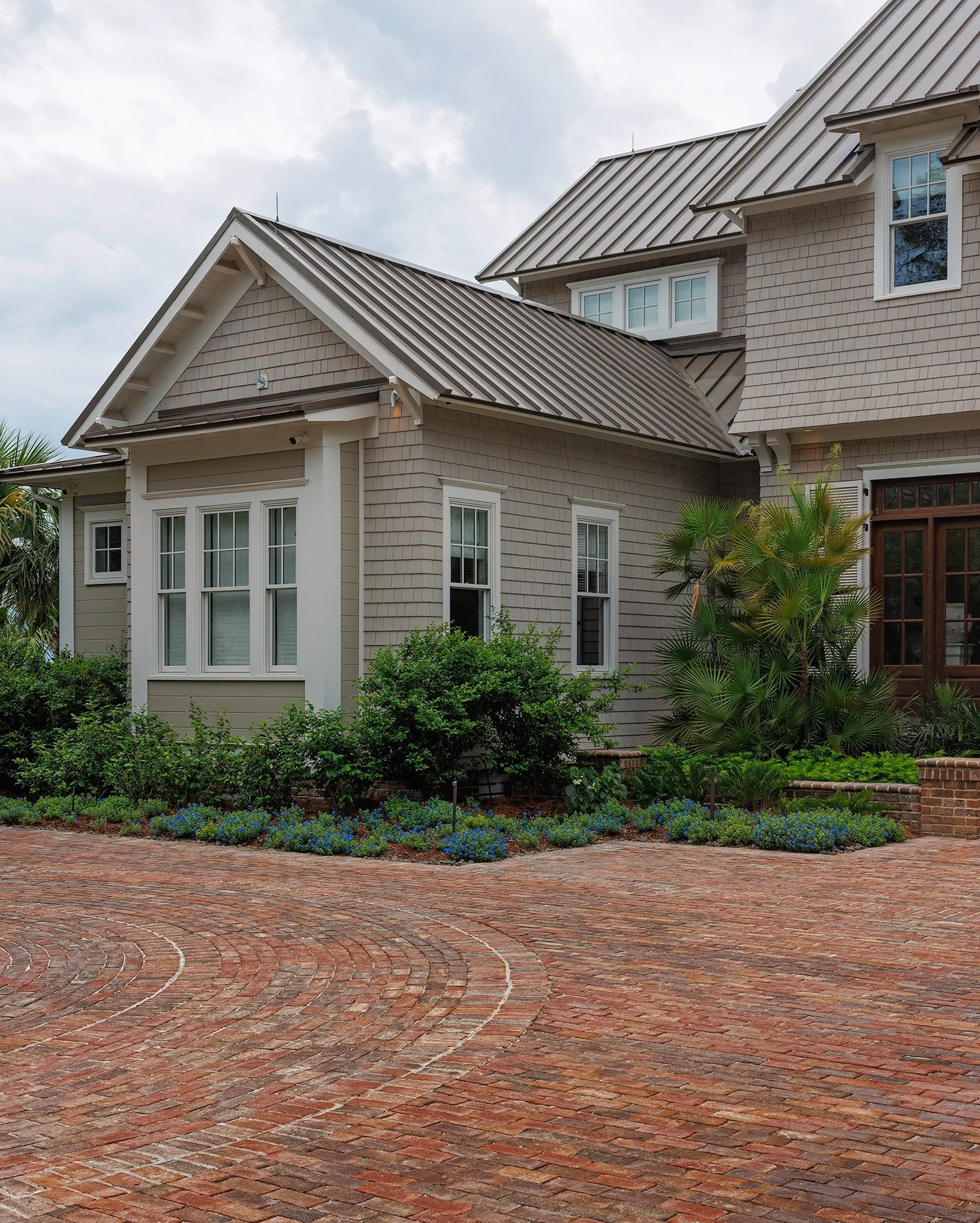 Exterior view of a house with beige siding, white trim, and a metal roof. There are multiple windows and a brick-paved driveway with landscaped bushes and plants in front.