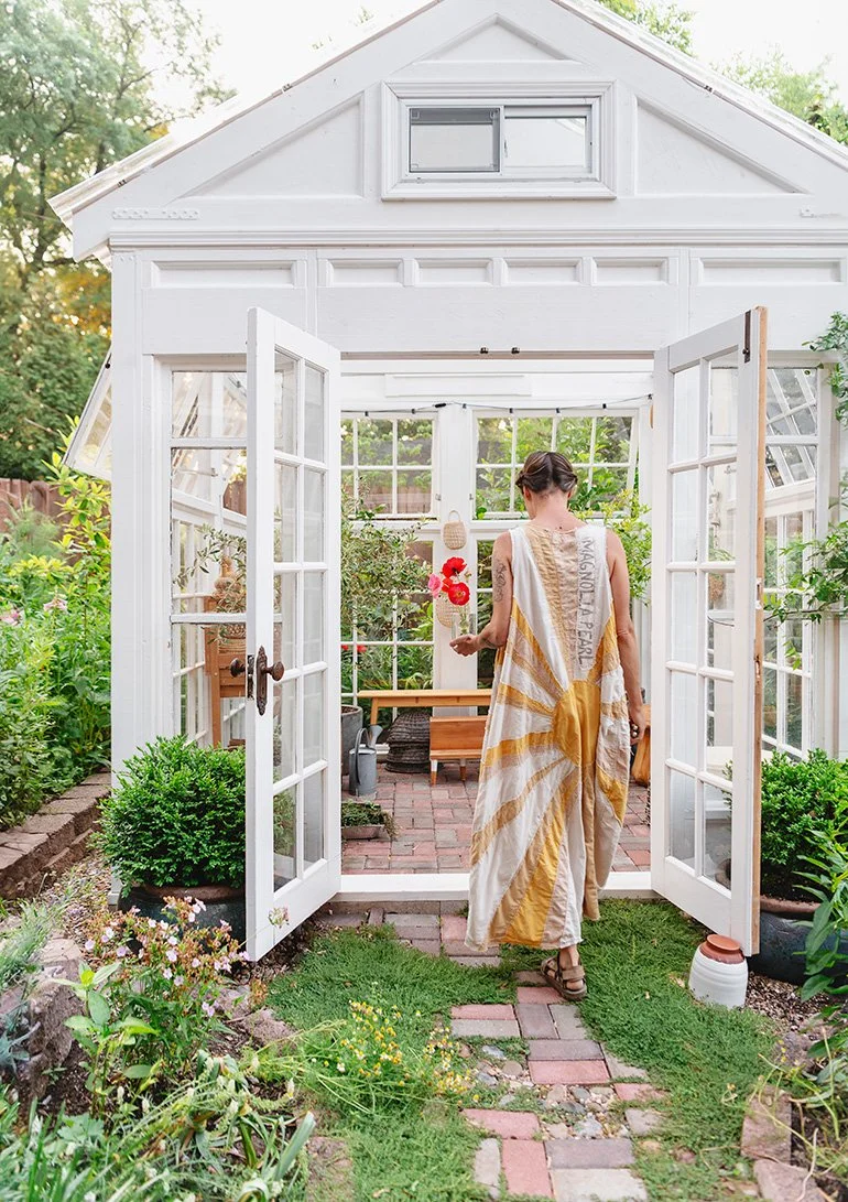 A woman in a white and mustard-colored dress walking into a white garden greenhouse with glass double doors, surrounded by lush green plants and flowers.