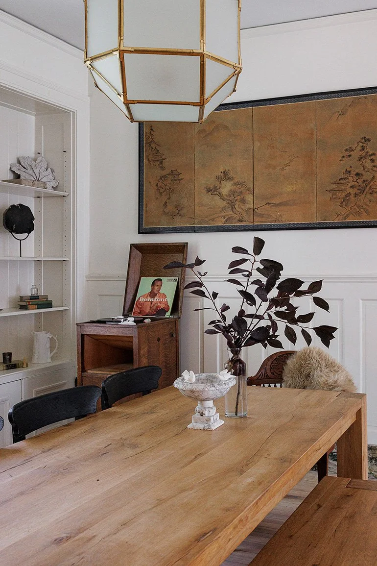 A dining room with a wooden table, a vase with dark leaves, a decorative pedestal with a white sculpture, and a fur-covered cushion on a wooden chair. A large wall hanging with a landscape scene is on the wall, and a vintage record player with a fram