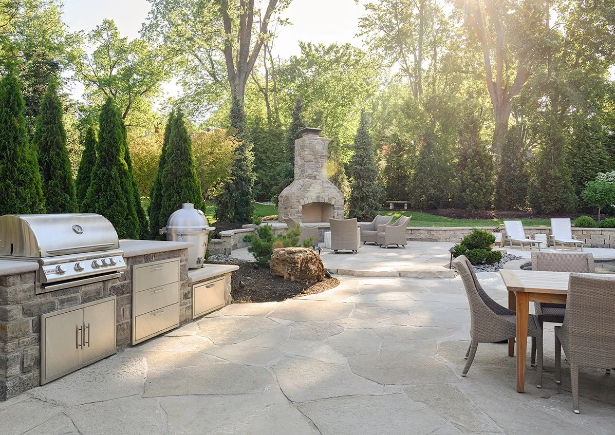 Outdoor patio with stainless steel grill, wood dining table, wicker chairs, stone fireplace, lounge chairs, lush green trees, and sunlight filtering through the trees.