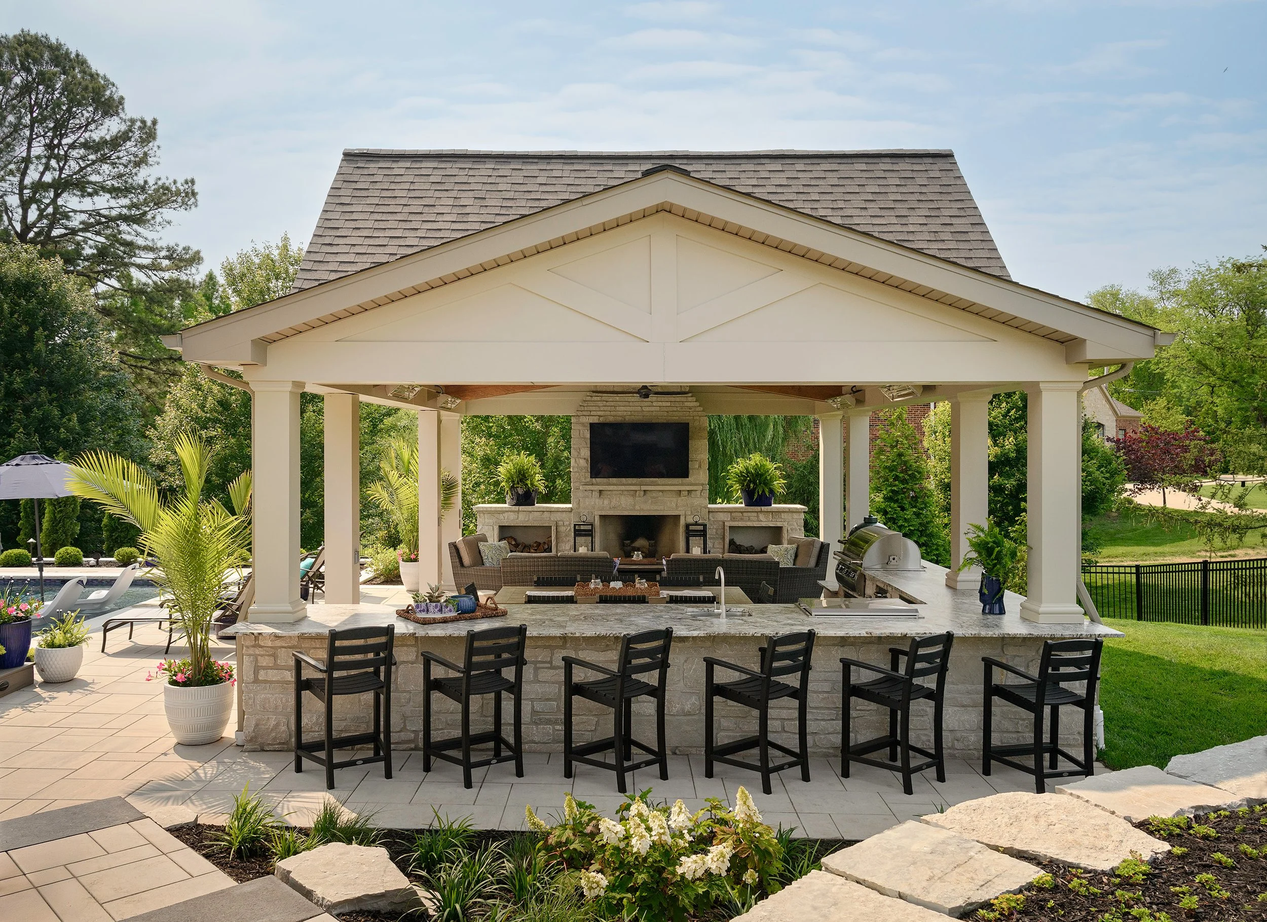 An outdoor kitchen and lounge area with a bar, seating, and a fireplace with a TV above, surrounded by greenery and trees.