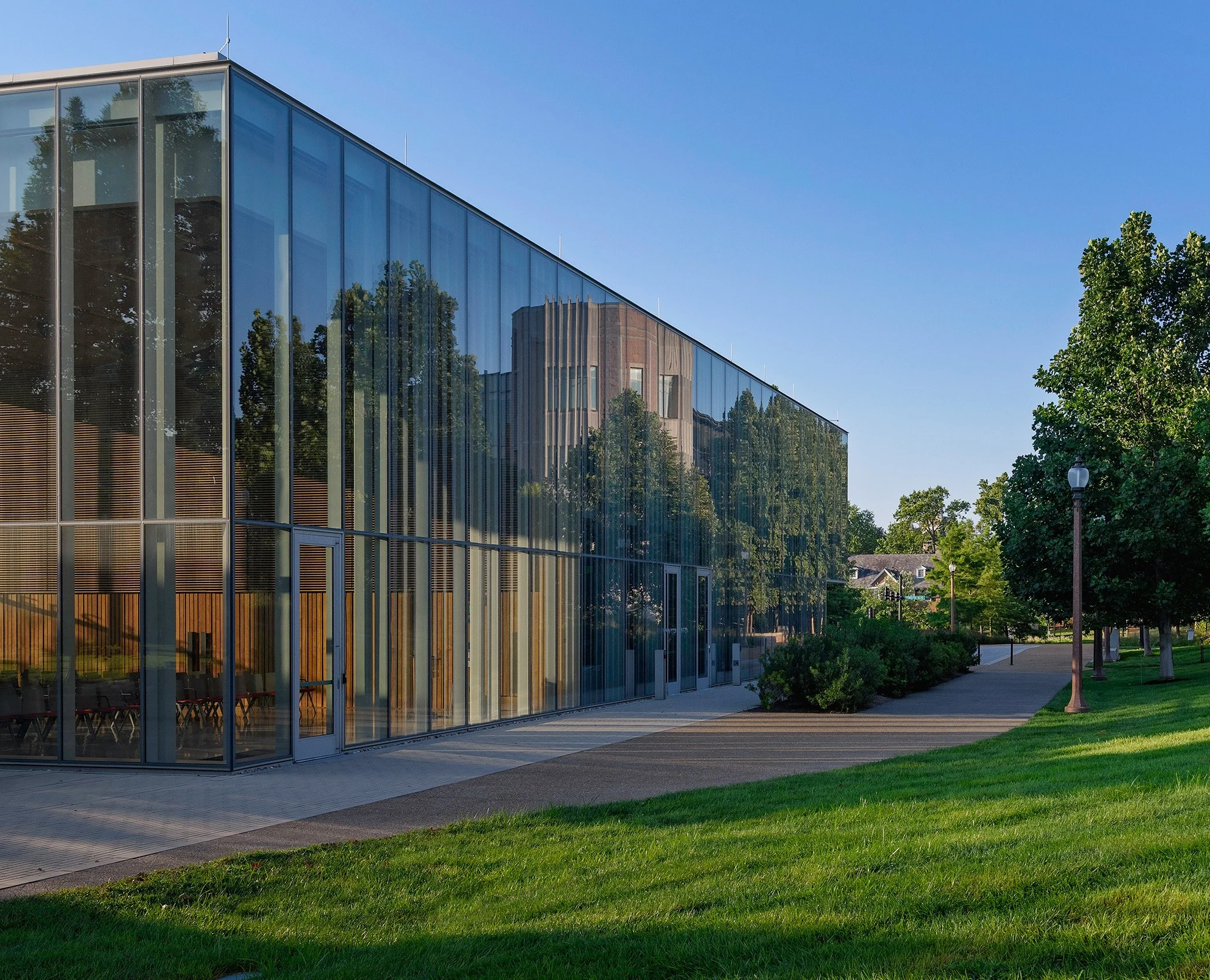 Modern glass building with trees and a pathway outside on a sunny day.