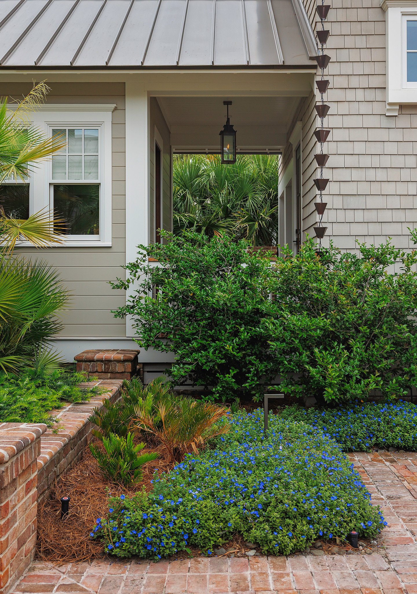 Front porch with brick pathway, green shrubs, blue flowering plants, and a house with beige siding, white window trim, and metal roof.