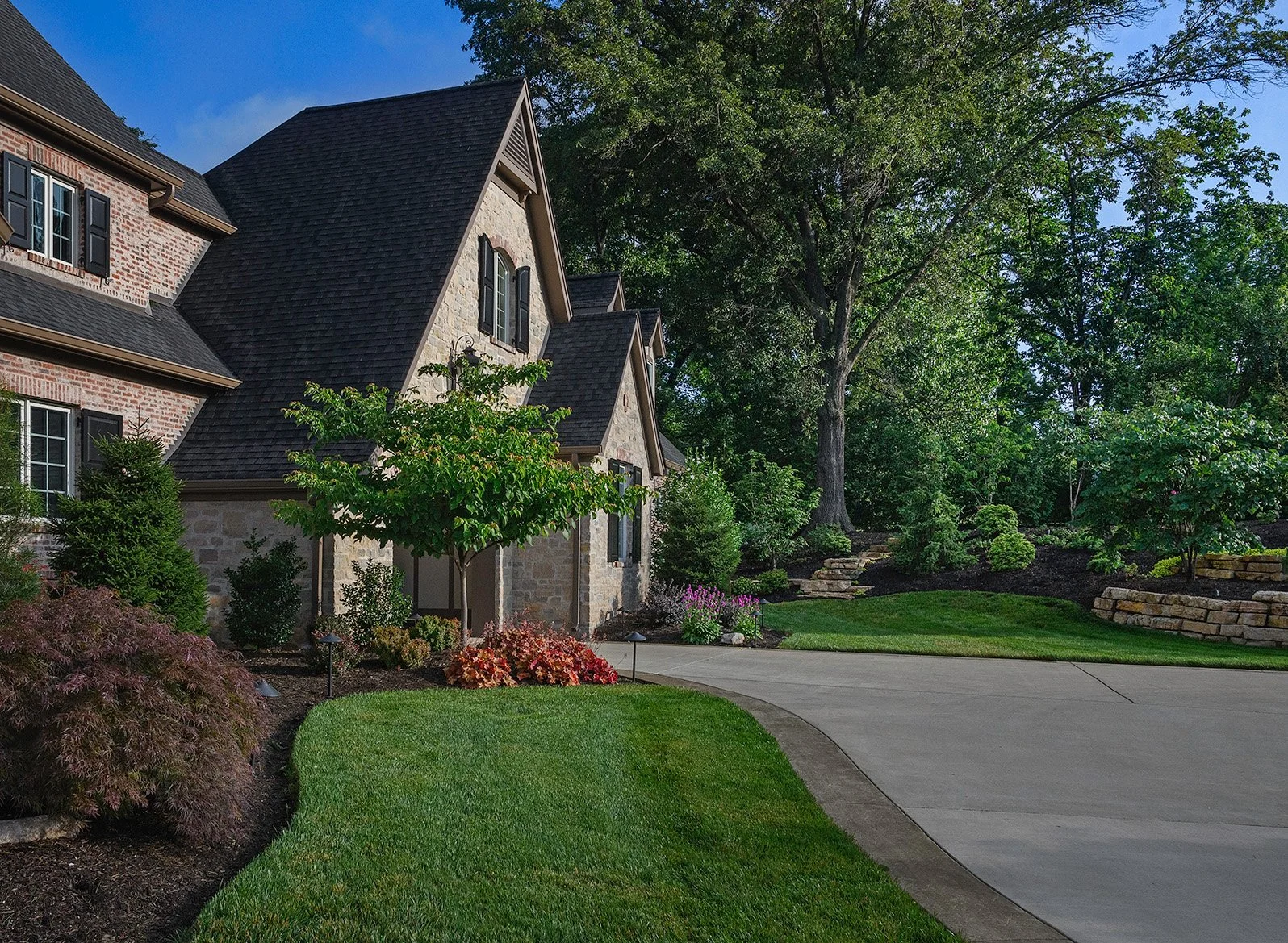 Landscape design photography of a front yard of a house with a driveway, lush green grass, trees, and landscaped bushes and flowers.