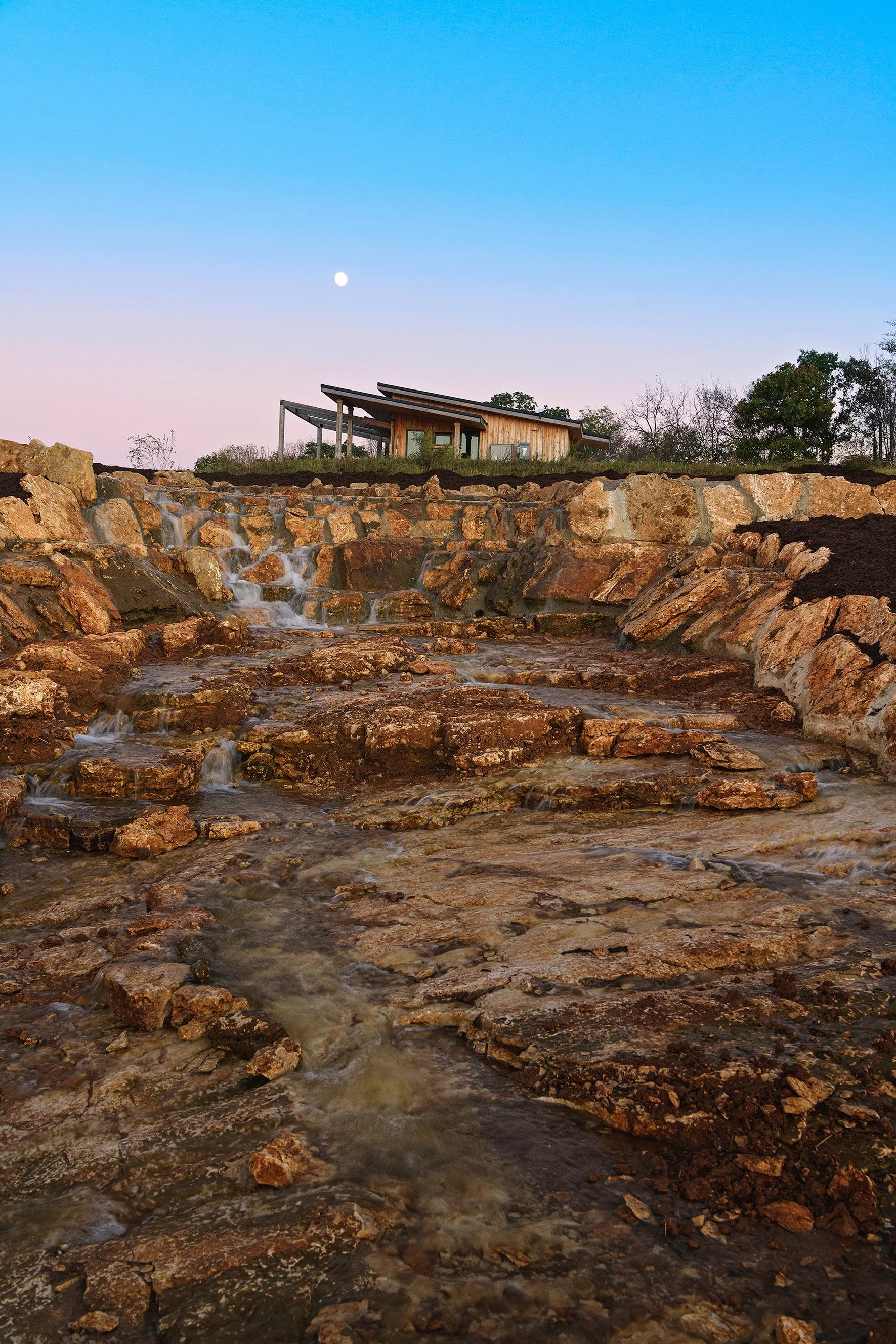 A small stream of water flowing over rocks in front of a modern wooden house on a hill, with a clear sky and the moon visible.