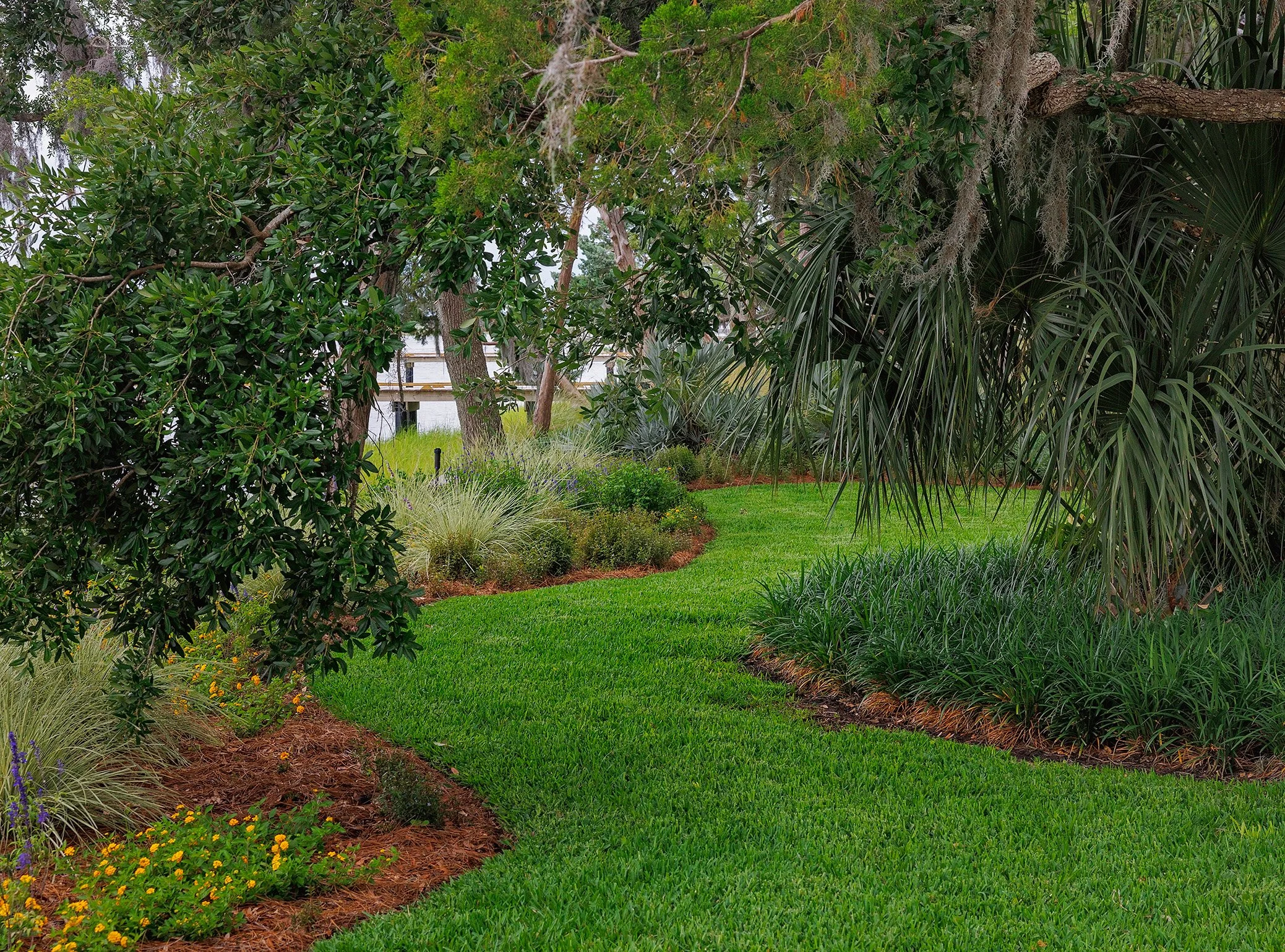 Lush green garden with well-maintained lawn, various shrubs, and tall trees. A wooden deck and water body are visible in the background.