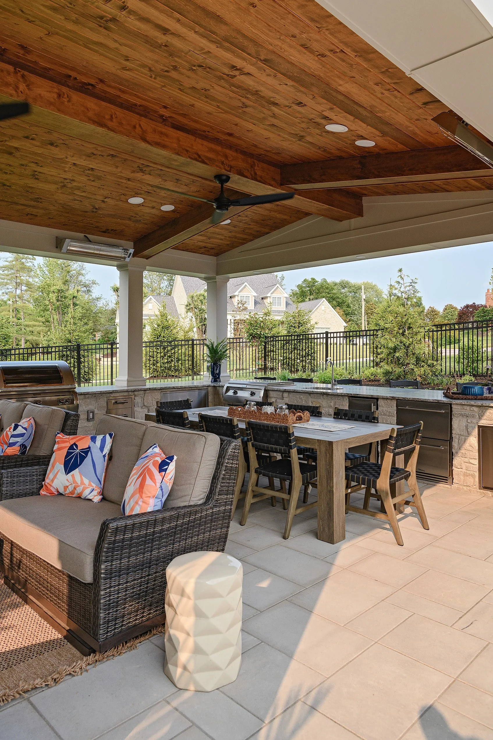 Outdoor covered patio with wicker sofa, colorful pillows, dining table with chairs, outdoor kitchen, and ceiling fan, overlooking a green yard and houses in the background.