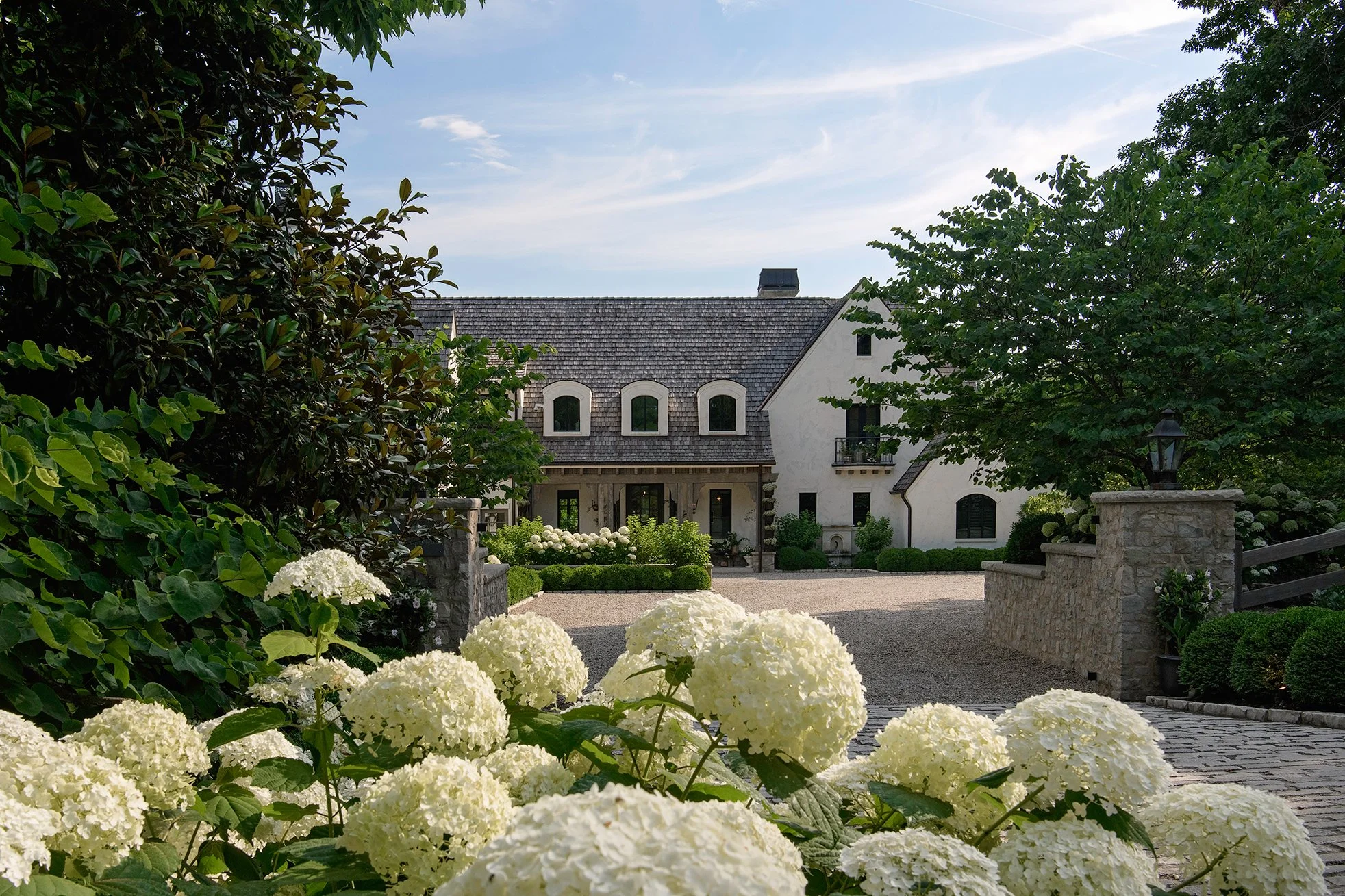 A white house with a dark roof surrounded by trees and white hydrangea flowers in the foreground, with a gravel driveway and decorative stone walls.