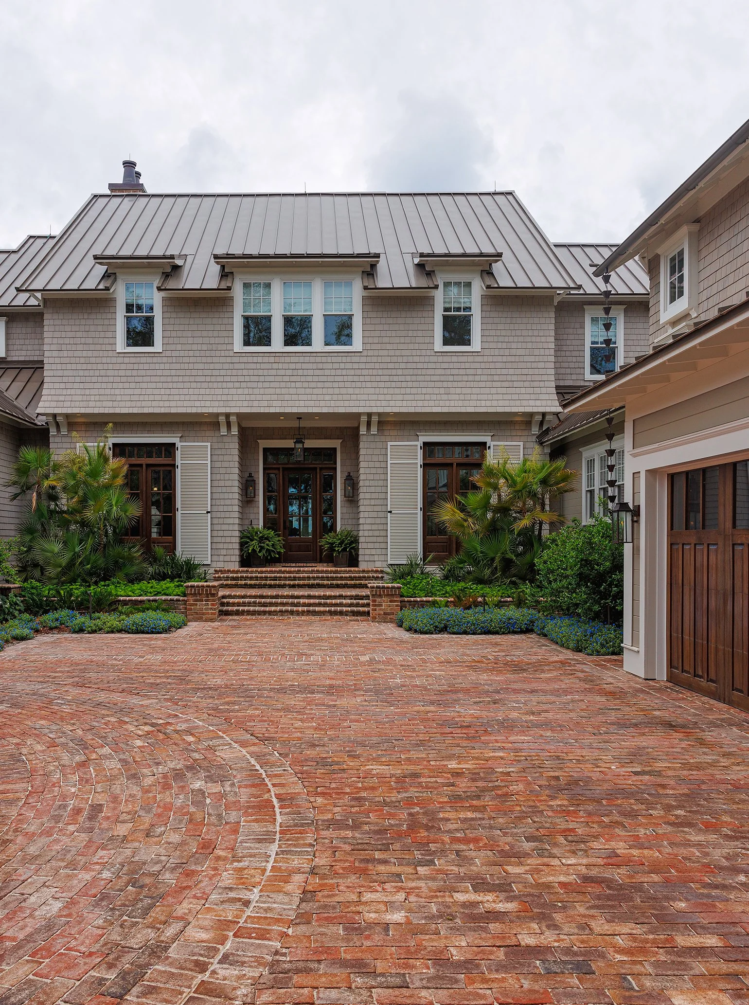 Front view of a large, two-story house with a brick driveway, lush plants, and trees, and a cloudy sky.