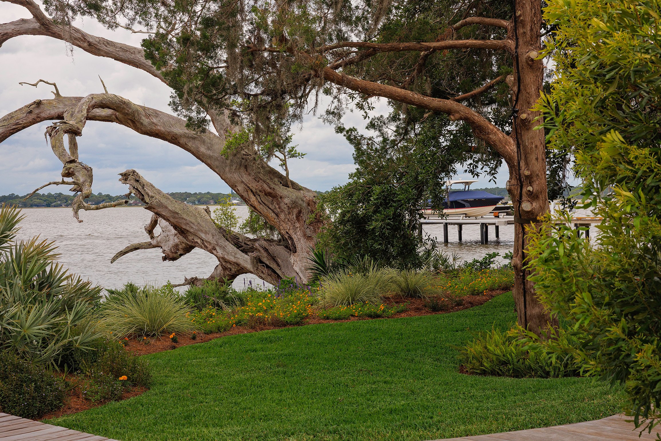 Lush garden with green grass, various plants, and a large tree by the water, with a boat docked on a pier in the background.