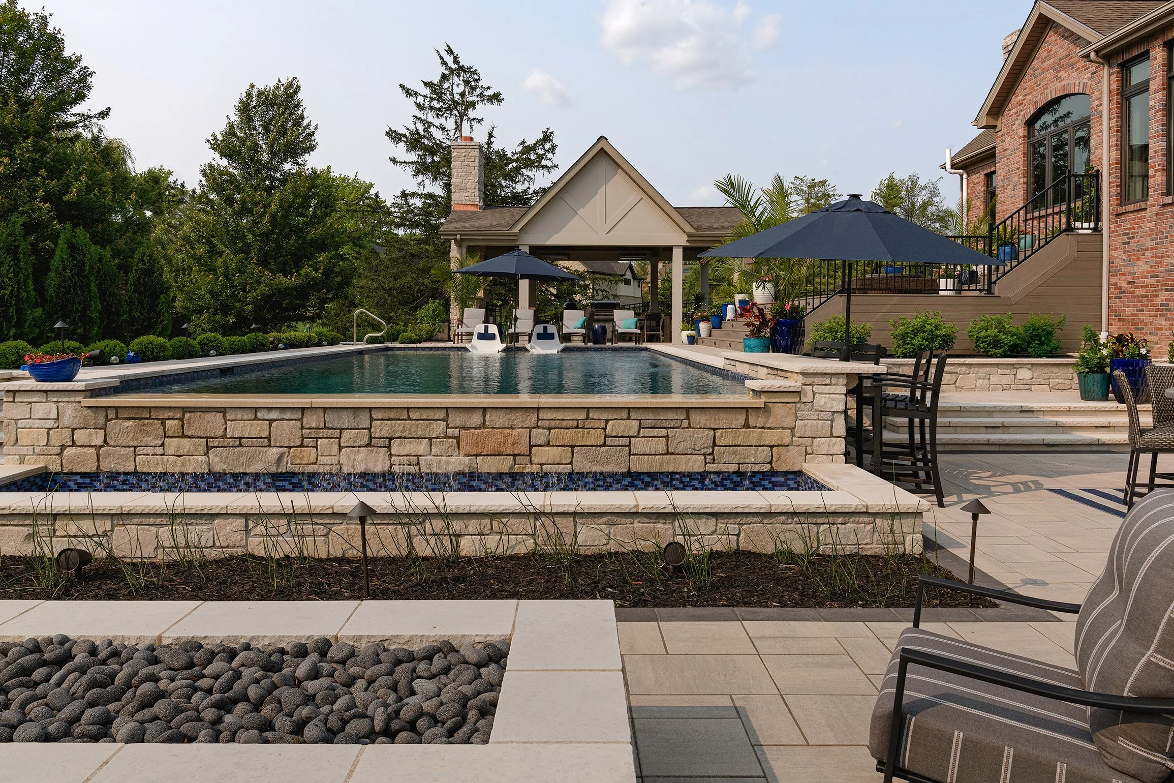 Backyard with in-ground pool, patio furniture with umbrellas, landscaped garden beds, and a brick house in the background.