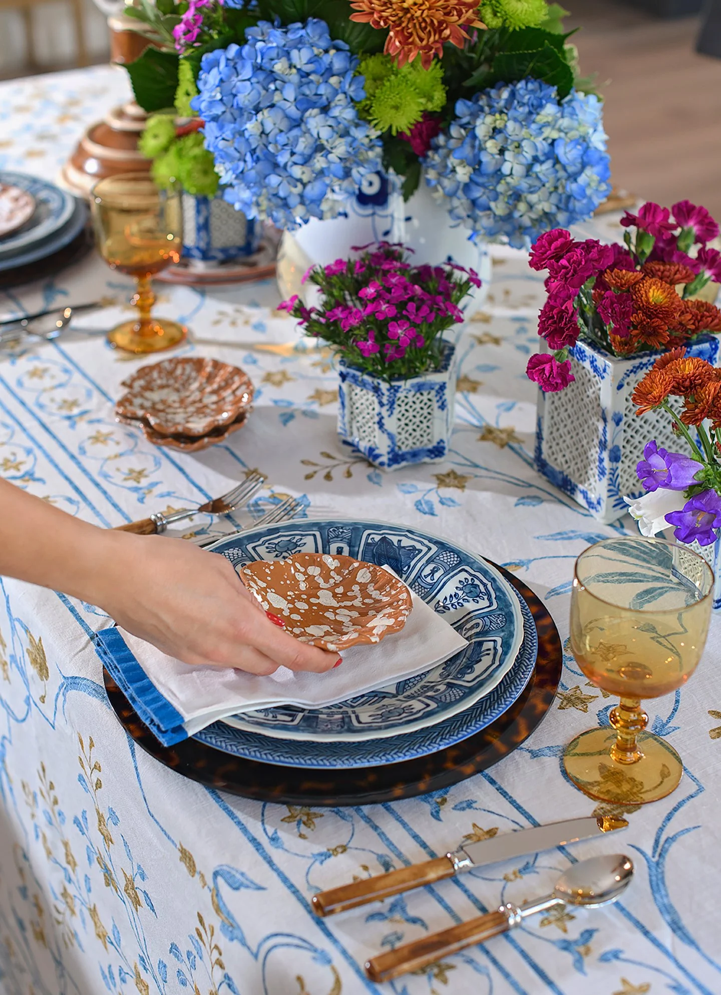 A table set with blue and white patterned plates, gold-rimmed glasses, and copper utensils. There are floral centerpieces with blue, pink, orange, and purple flowers in decorative white and blue vases, on a tablecloth with a floral and star pattern.