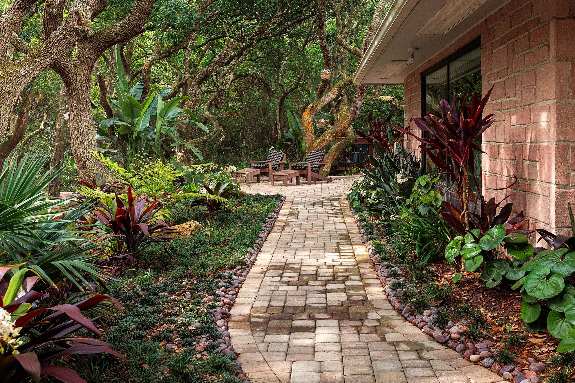 A garden pathway made of bricks and stones leading to a seating area with chairs, surrounded by lush green plants and trees next to a brick house.