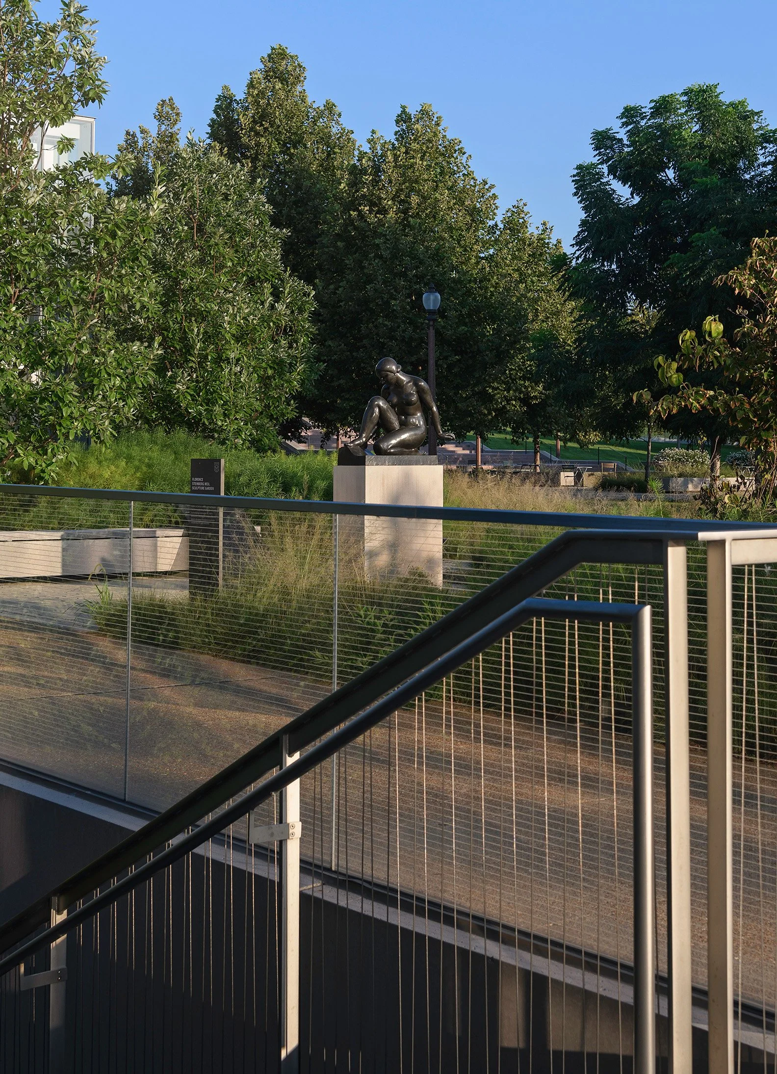 Sculpture of a seated woman outdoors in a park with trees, lamp post, and a sign visible in the background.