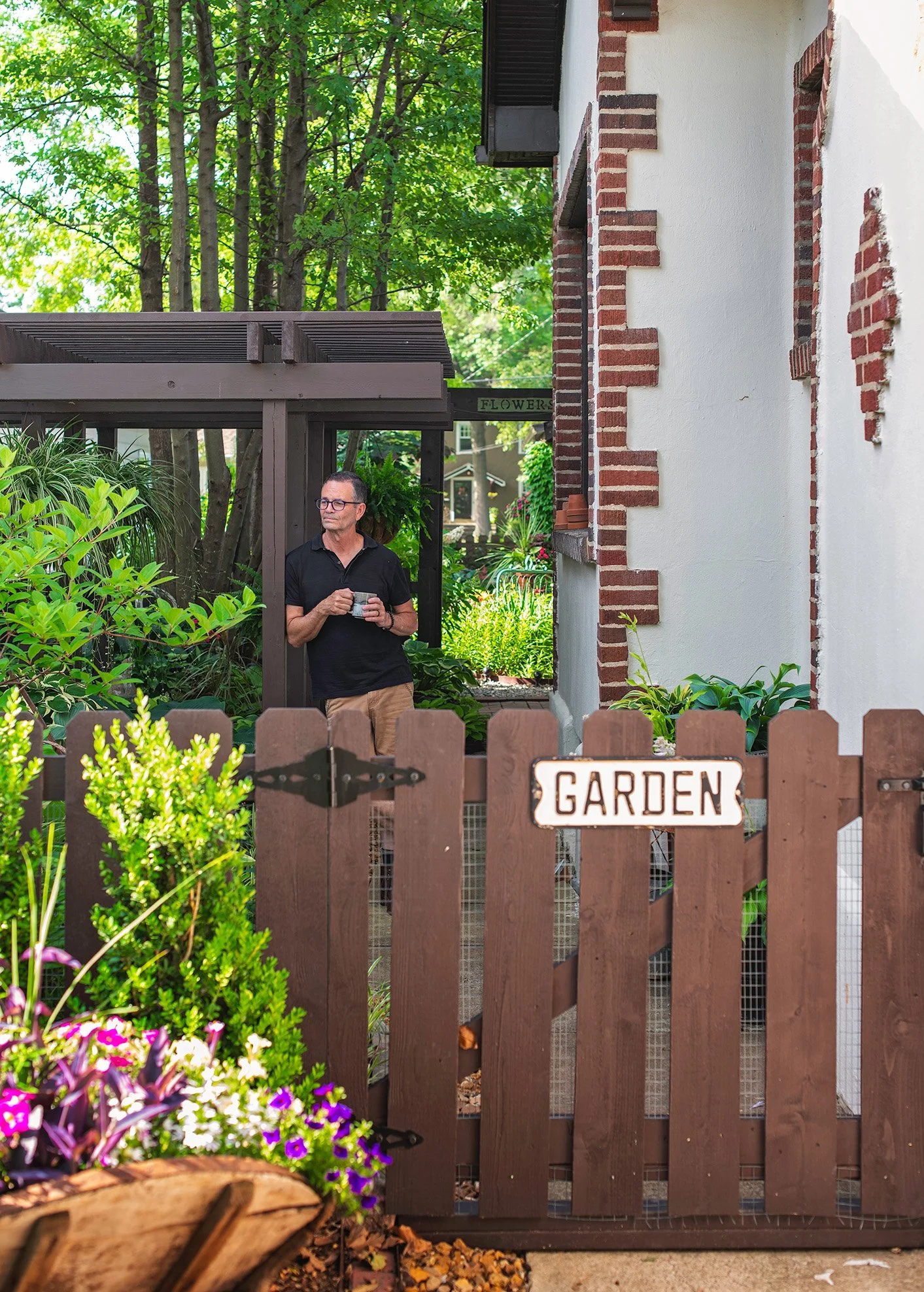 A man standing in a garden, holding a cup, behind a wooden gate labeled 'GARDEN.' The garden is lush with green plants and flowers, with a house wall in the background.