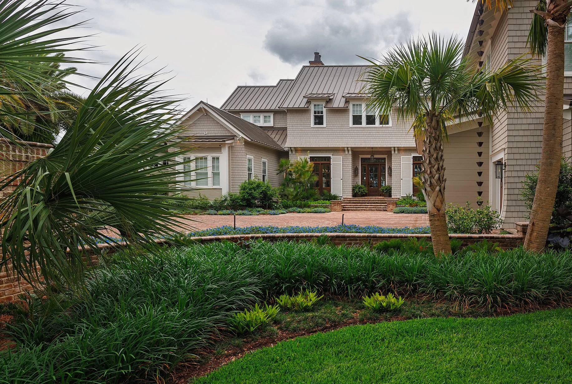A large house with a metal roof and a landscaped front yard featuring various green plants, palm trees, and a brick walkway.