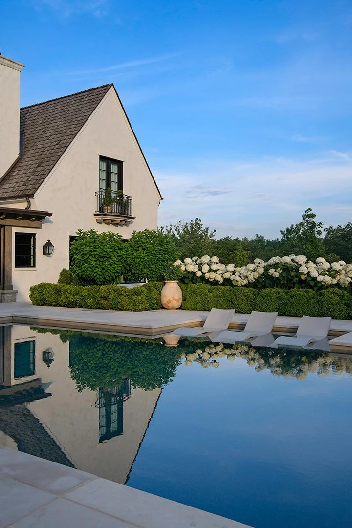 A backyard with a swimming pool, lounge chairs, and lush bushes and flowers, with a house featuring a small balcony against a blue sky.